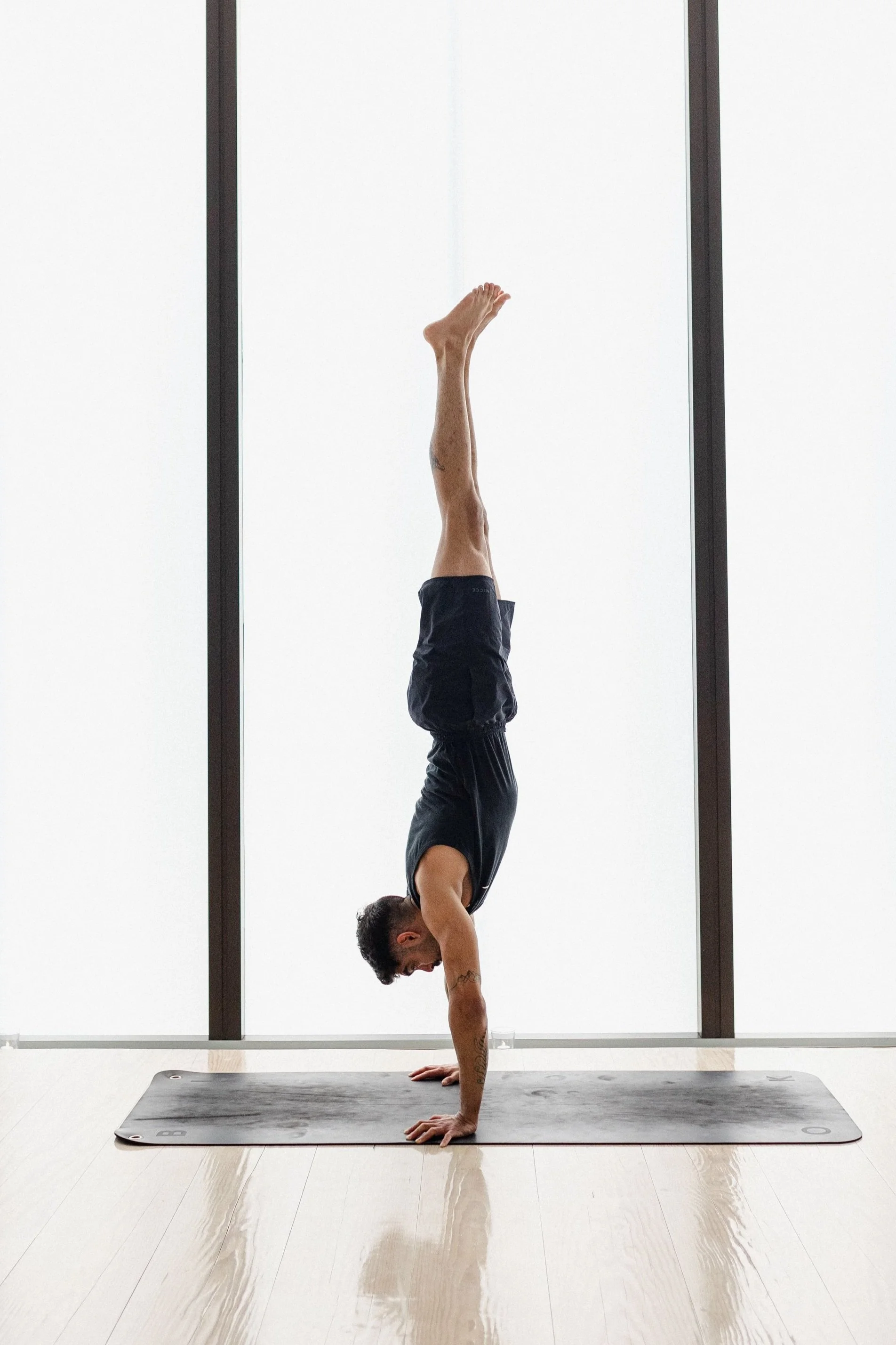 A man practicing handstand yoga pose on a gray mat in front of large windows in a bright room.