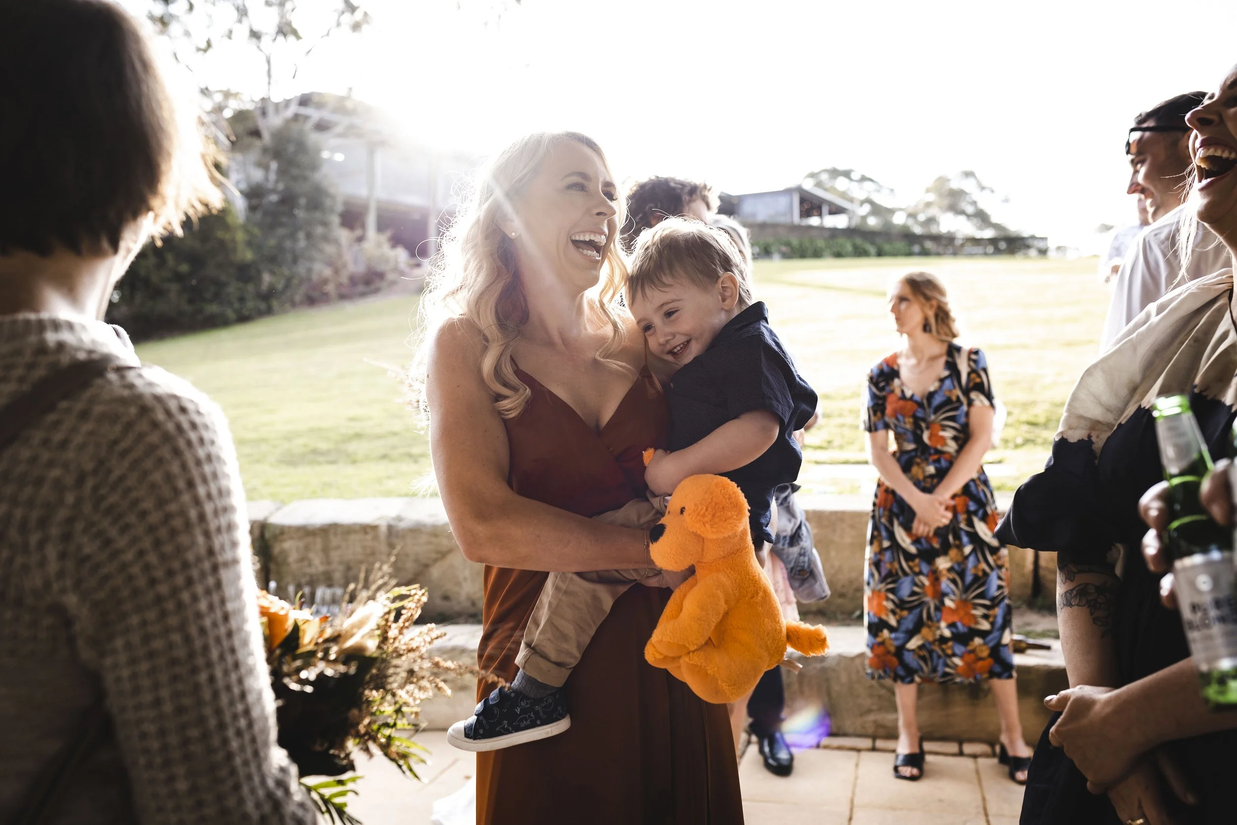 A woman holding a laughing young boy with a toy, surrounded by smiling people outdoors on a sunny day.