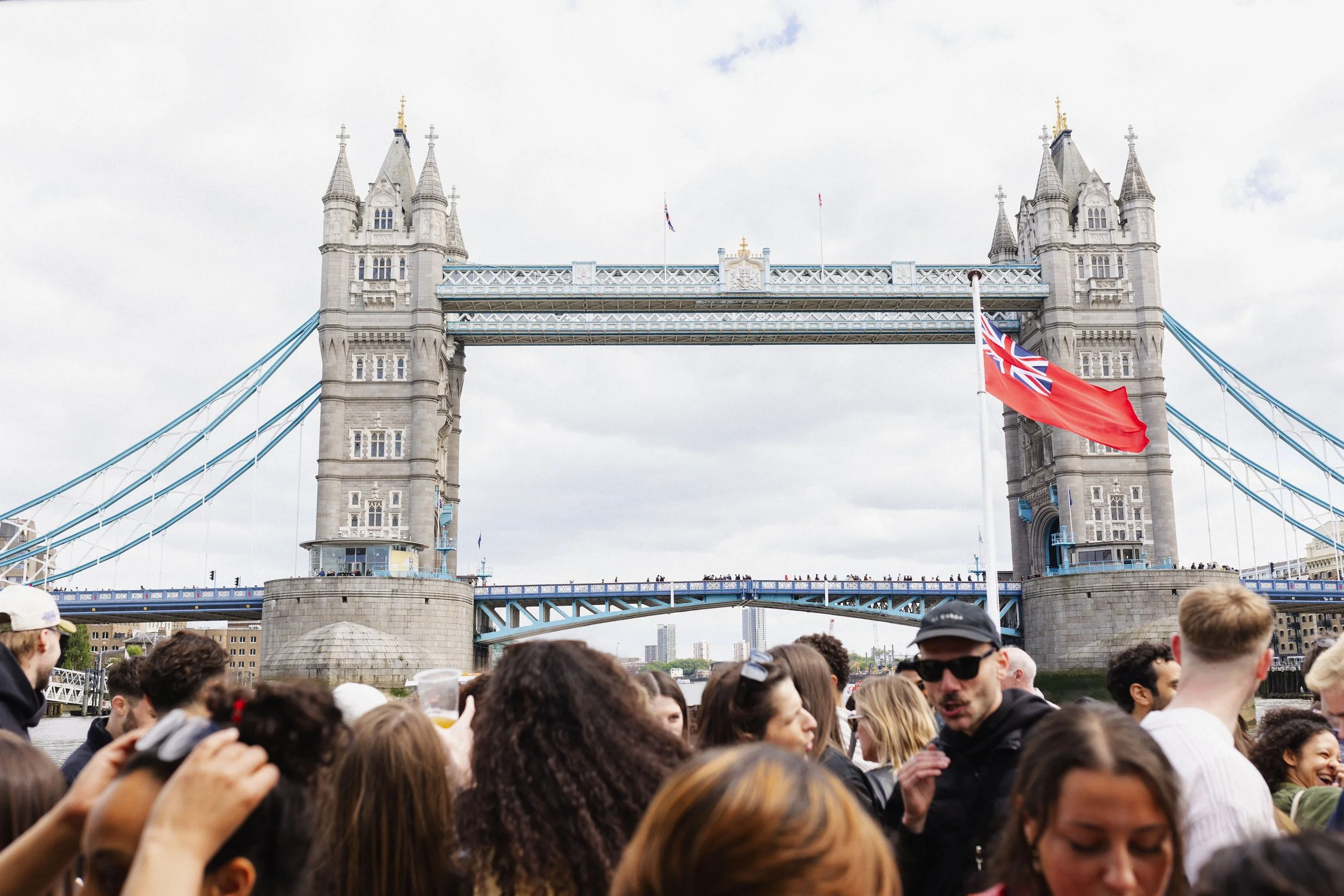 Crowd of people gathered near Tower Bridge in London, with the bridge's towers and suspension cables visible in the background, and an English flag flying in the foreground.