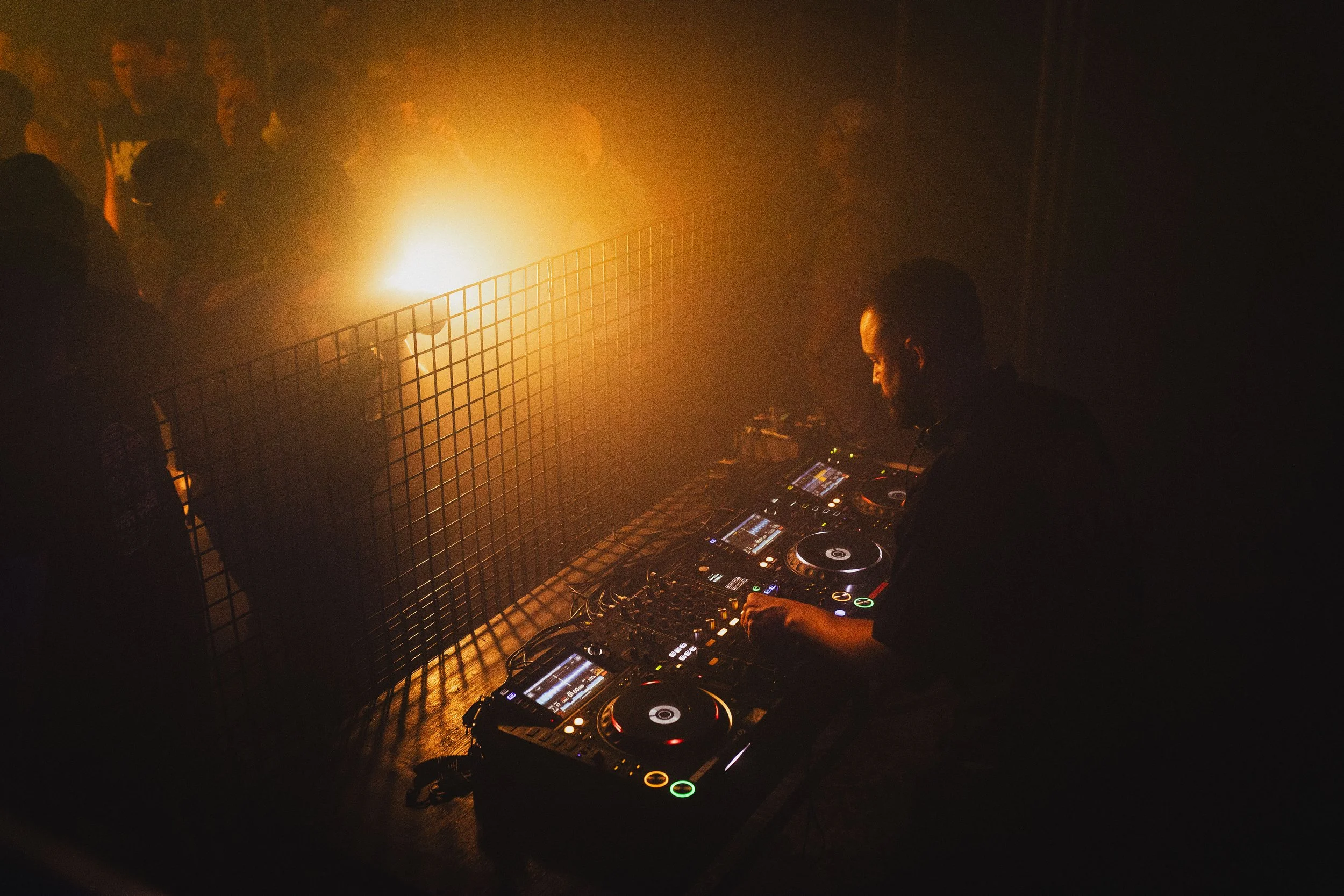 A DJ performing with turntables and audio equipment in a dimly lit club or party setting, with a crowd of people dancing and a bright light illuminating the scene from behind.