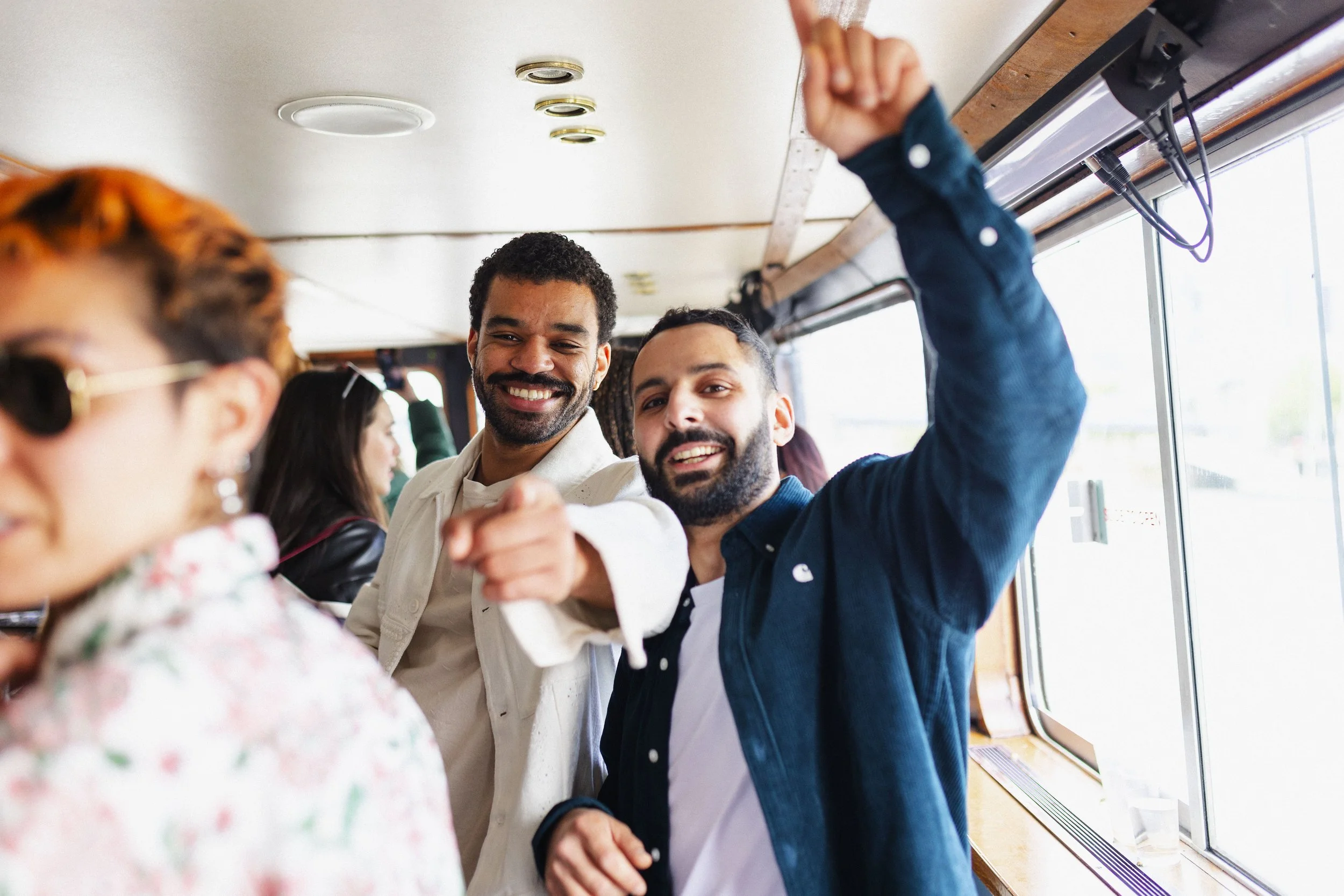 Two smiling men pointing towards the camera on a bus, with a woman in sunglasses in the foreground.