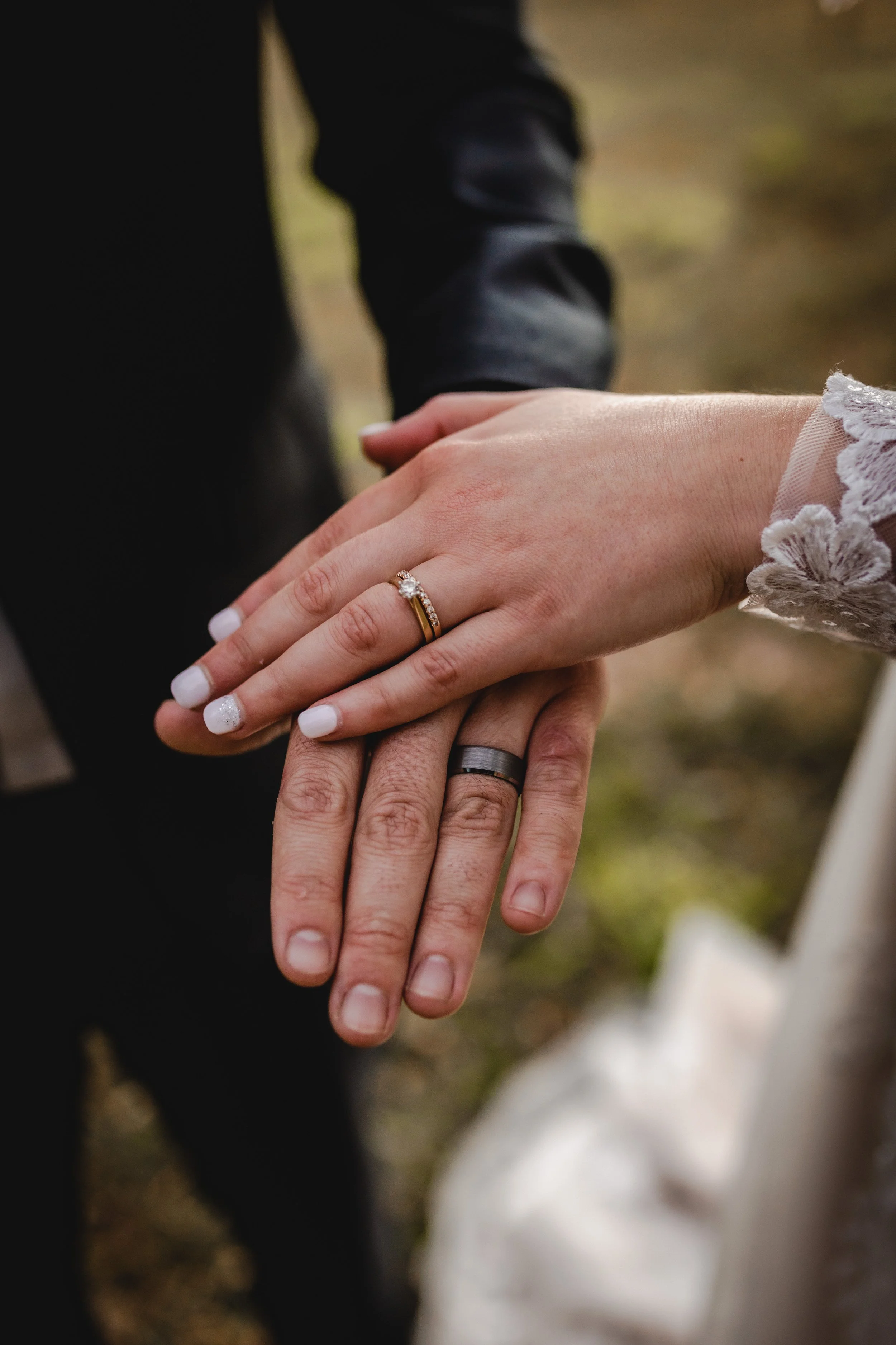 Close-up of a bride and groom's hands showing wedding rings, with the bride's hand on top. The bride's ring features a diamond and is white gold, while the groom's ring is a dark metal. The bride is wearing a lace wedding dress.