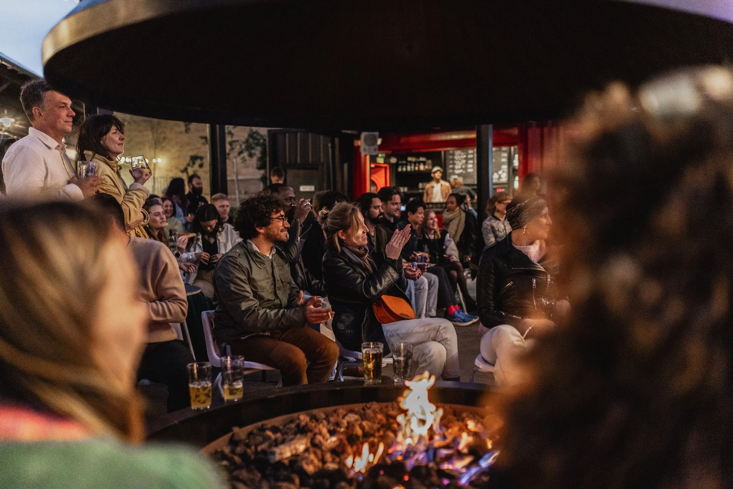 An outdoor evening gathering of people watching a performance or event with some seated and some standing, around a fire pit.