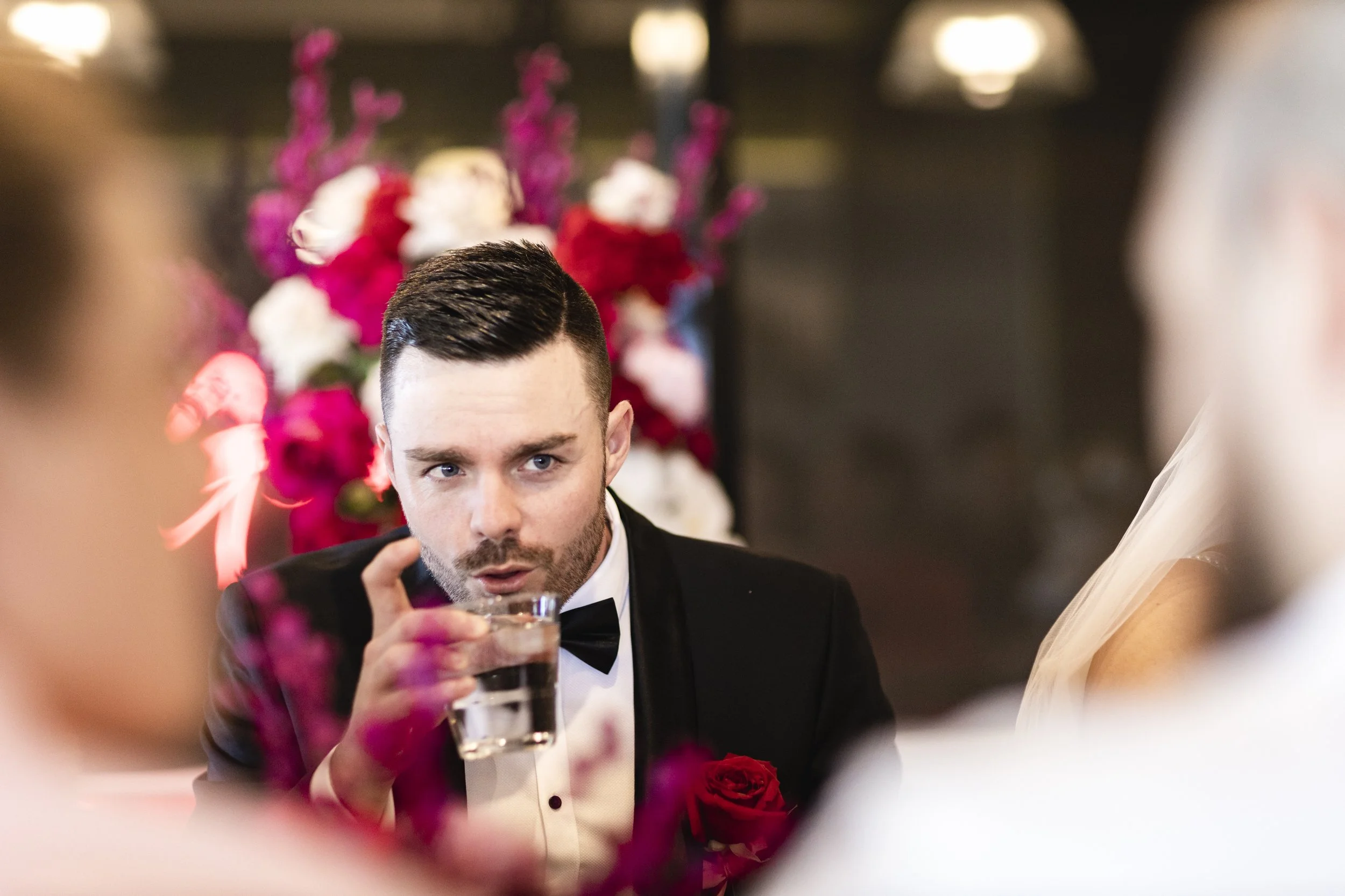 A man in a tuxedo with a bow tie is holding a glass of drink at a formal event, with a woman in a wedding dress and veil partly visible on the right, and blurred pink and white flowers in the background.