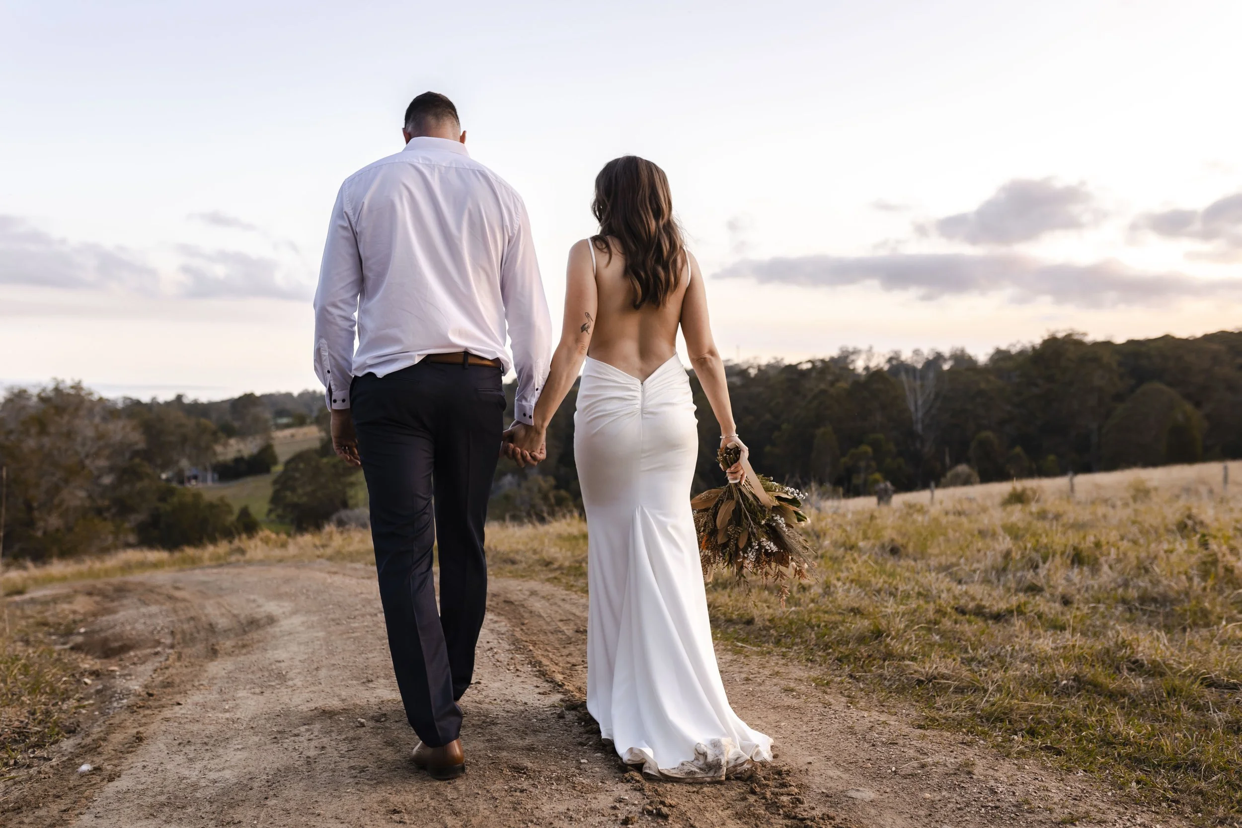 A couple holding hands walking away on a dirt path in a field at sunset, the woman wearing a white wedding dress and carrying a bouquet, and the man in a white shirt and dark pants.