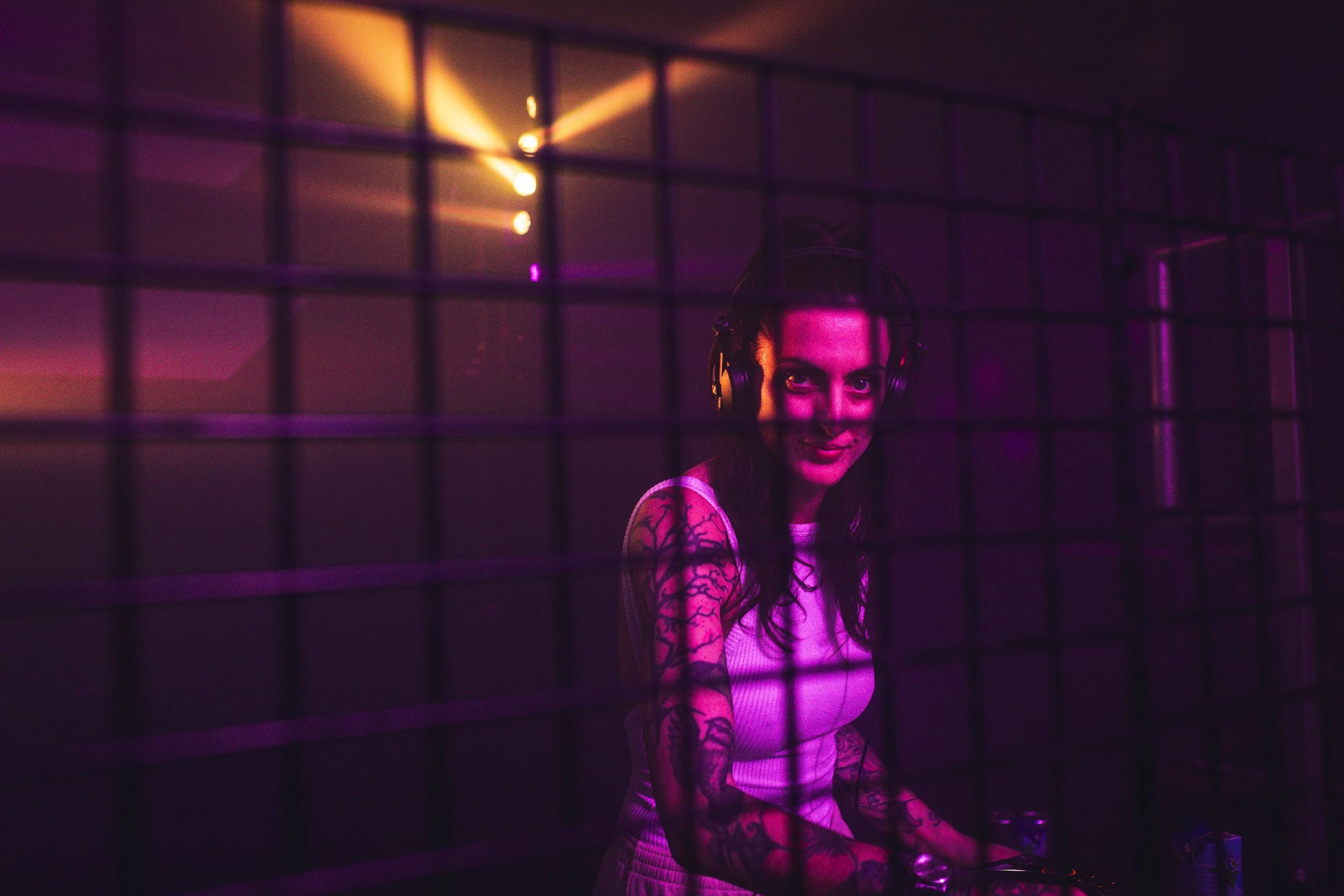 A woman with tattoos wearing headphones, smiling at a DJ booth illuminated with purple and pink lighting, behind a metal grid.