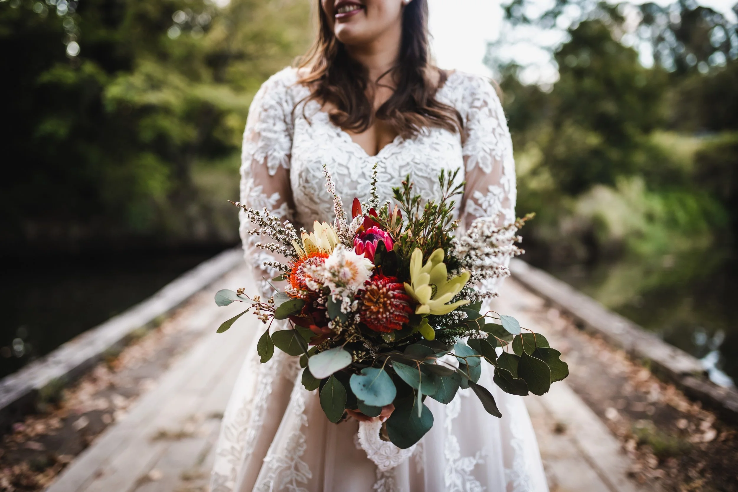 Woman in a lace wedding dress holding a colorful bouquet of flowers outdoors, standing on a wooden bridge with greenery in the background.