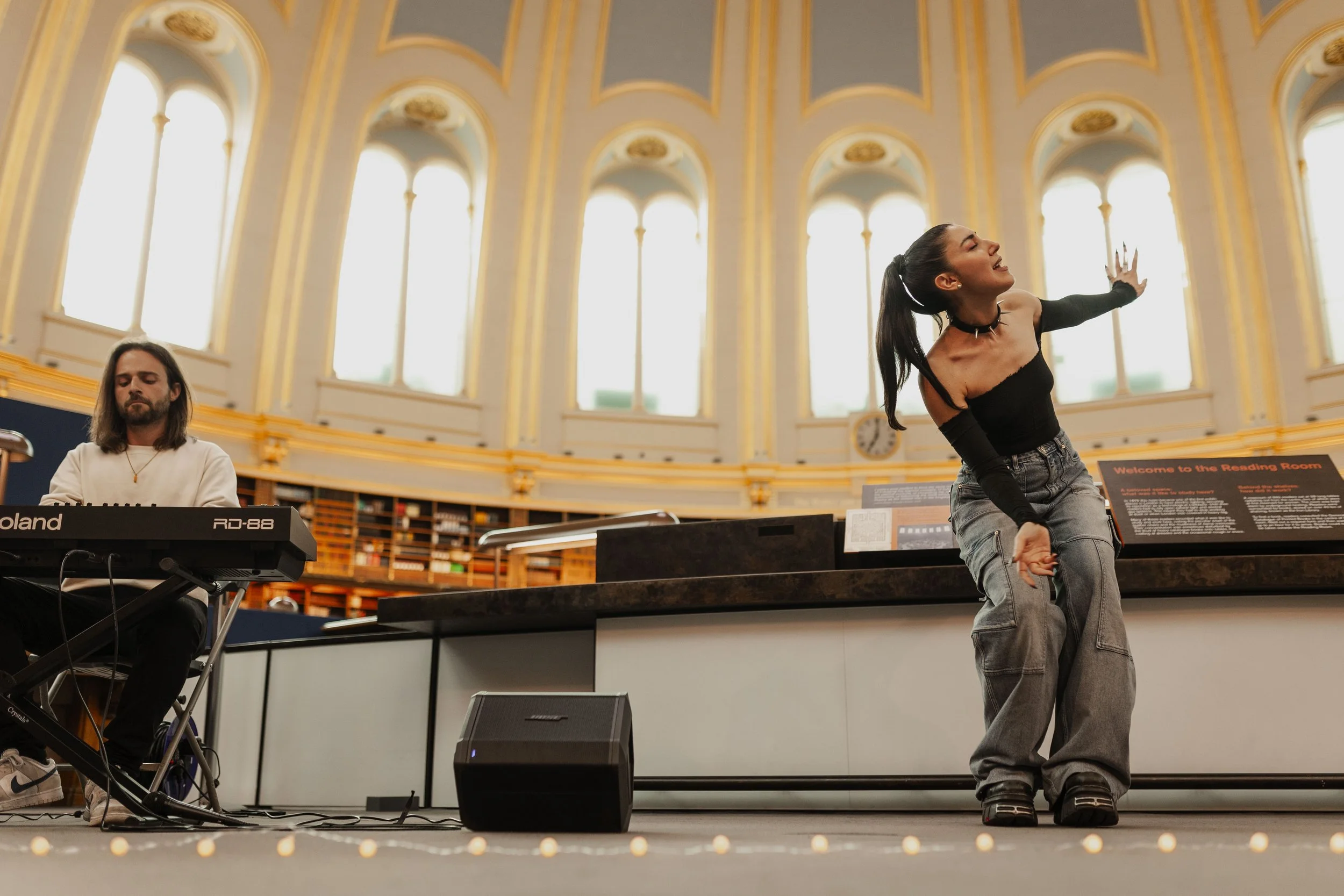 A woman performing a dance in front of a keyboard player in a large, ornate hall with tall windows.