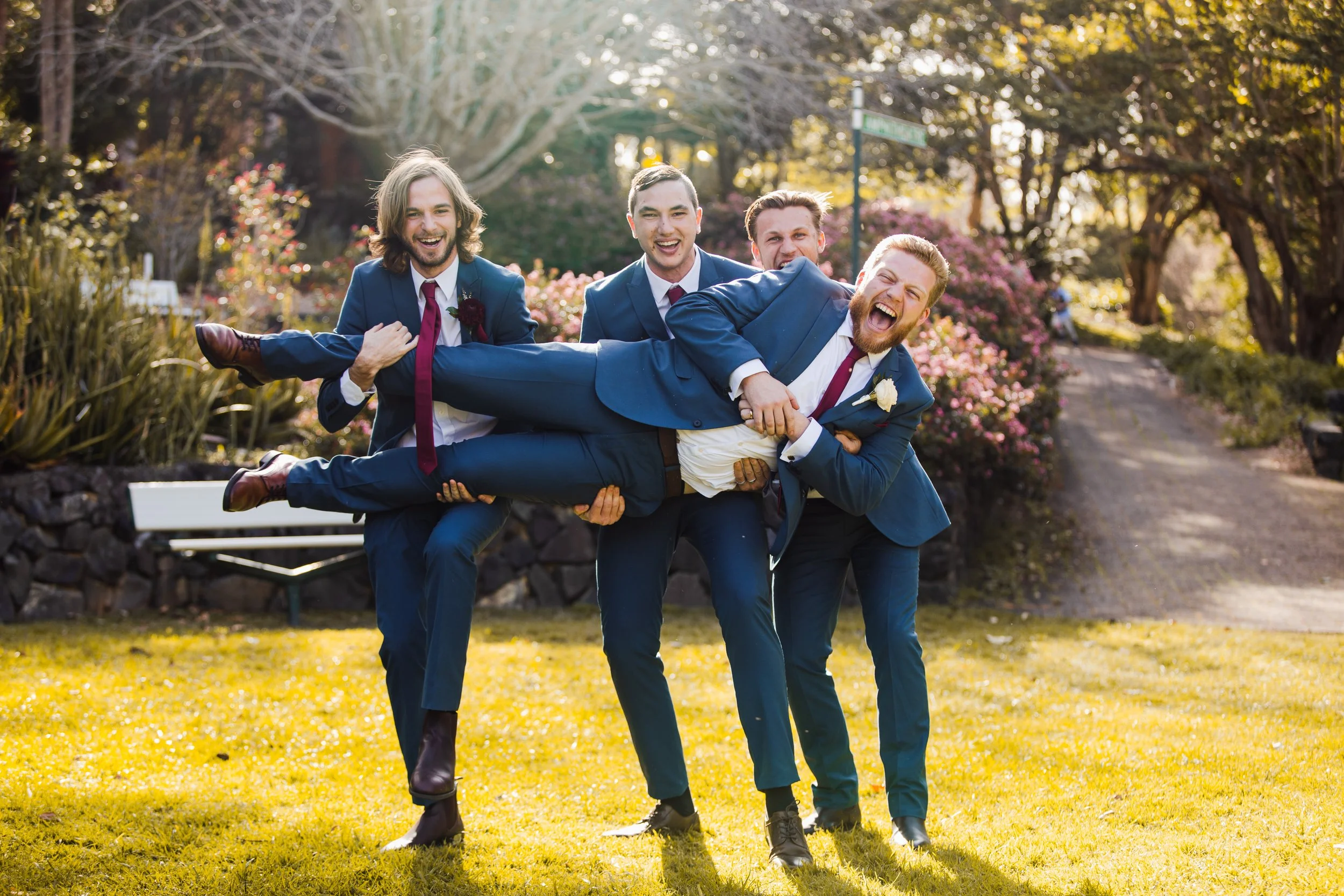 Four men in suits are outdoors, with three of them holding up a smiling man horizontally. The men are dressed in blue suits with ties, and the scene is bright with sunlight, surrounded by trees and pink flowering bushes.