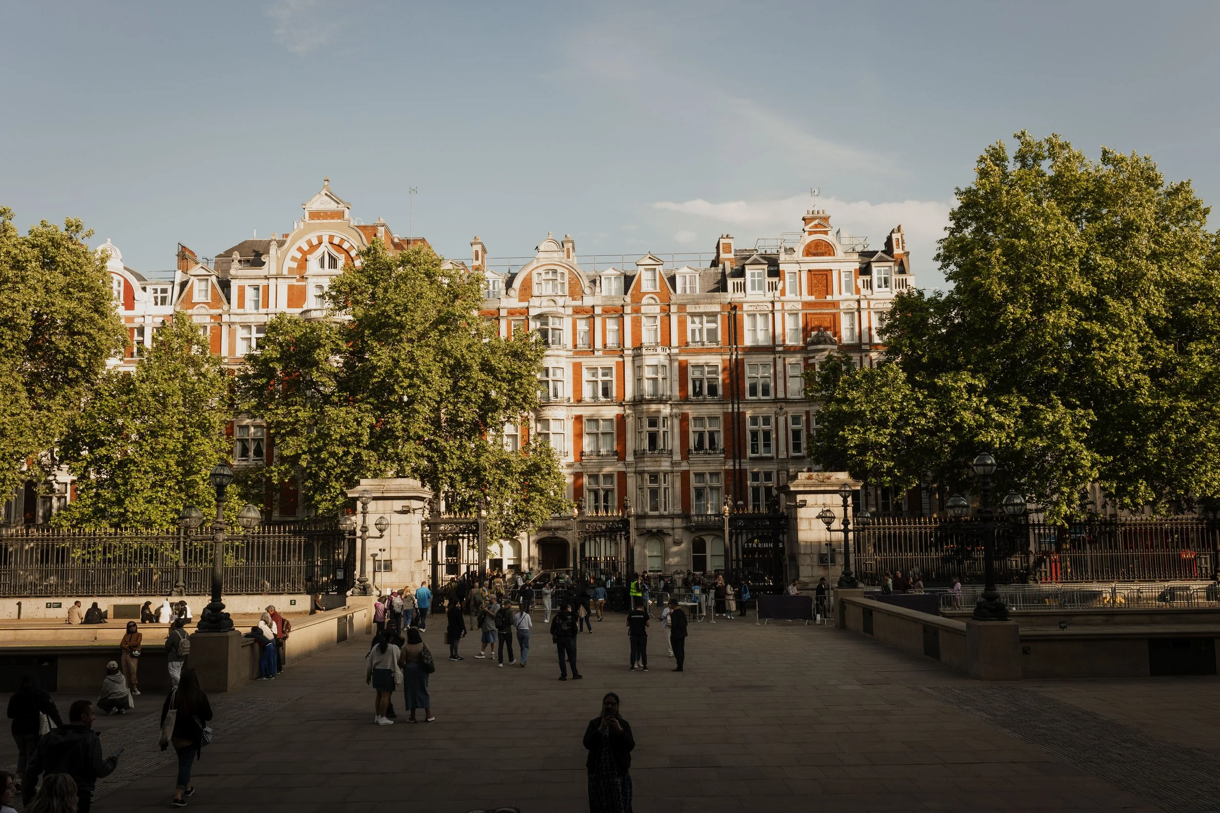 City square with people walking and sitting, ornate multi-story building with red brick and white accents, large trees, street lamps, and a black metal fence