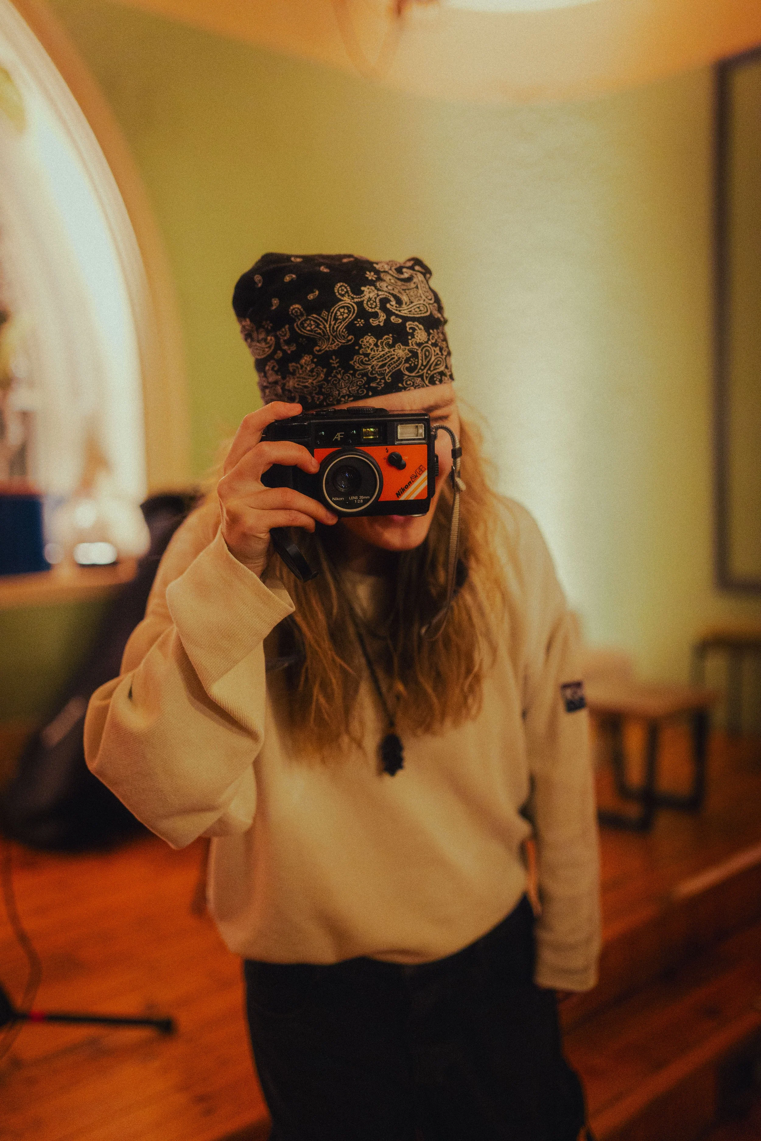 Person with long hair, wearing a bandana and beige sweatshirt, taking a photo with a vintage camera in a room with green walls, a wooden desk, and a backpack.
