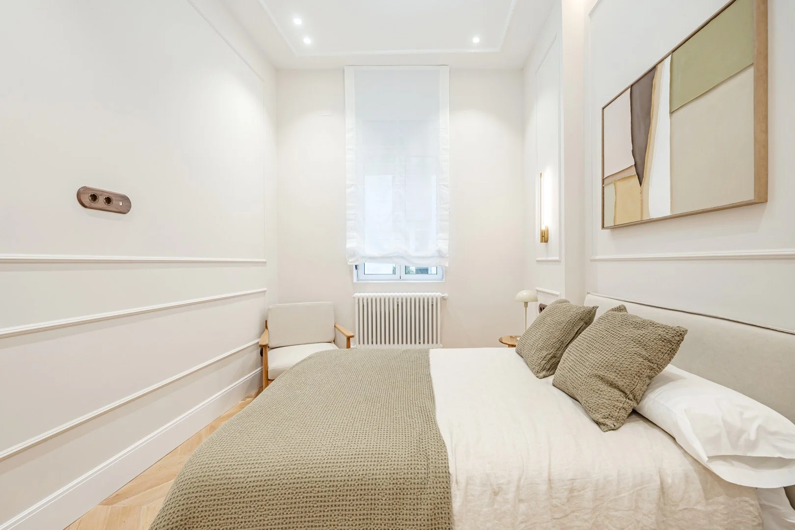 Minimalist bedroom with white walls, a window with sheer curtains, a bed with beige bedding and pillows, a small bedside table with a lamp, a white armchair, and abstract wall art.