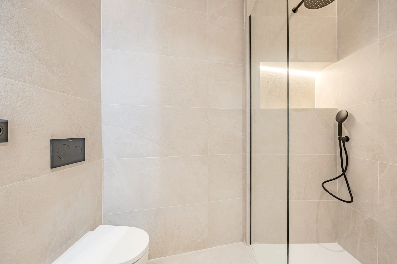Minimalist modern bathroom with beige tiled walls, a wall-mounted toilet, and a walk-in shower with a black handheld showerhead and a glass partition.