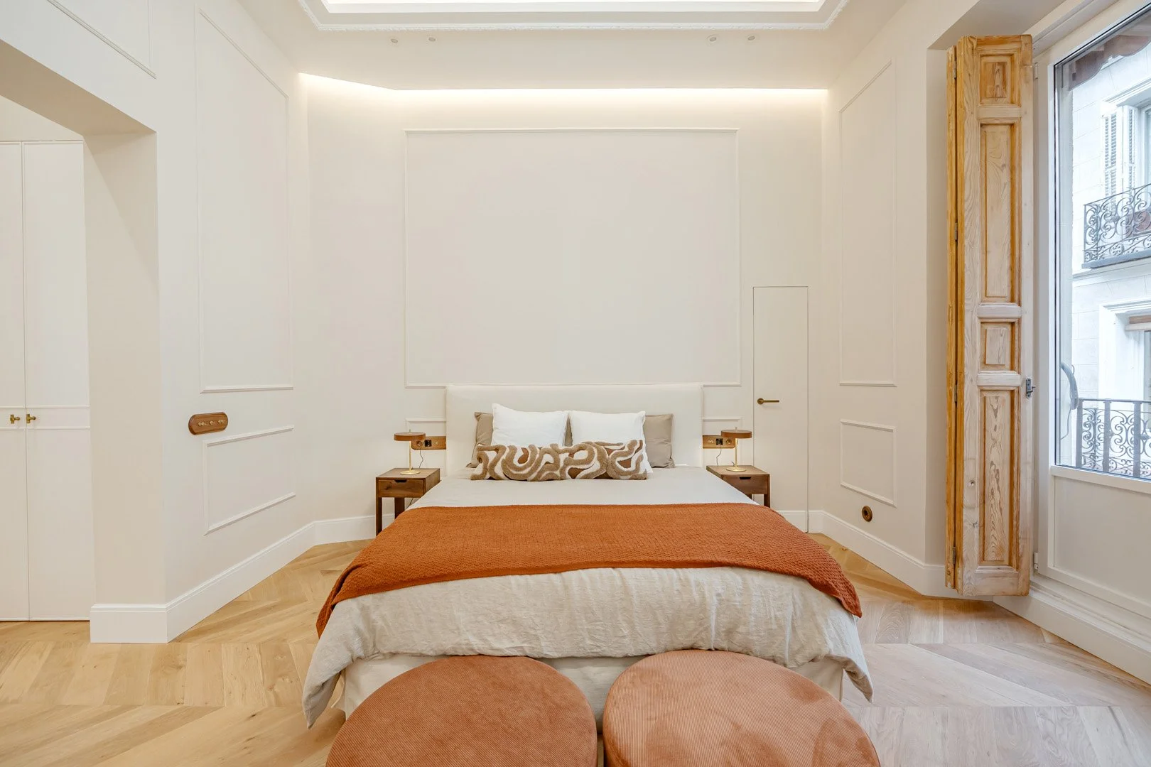 Modern bedroom with a large white bed, beige and white pillows, rust-colored throw, and two brown nightstands with lamps, wooden floor, and a window with a wooden shutter.