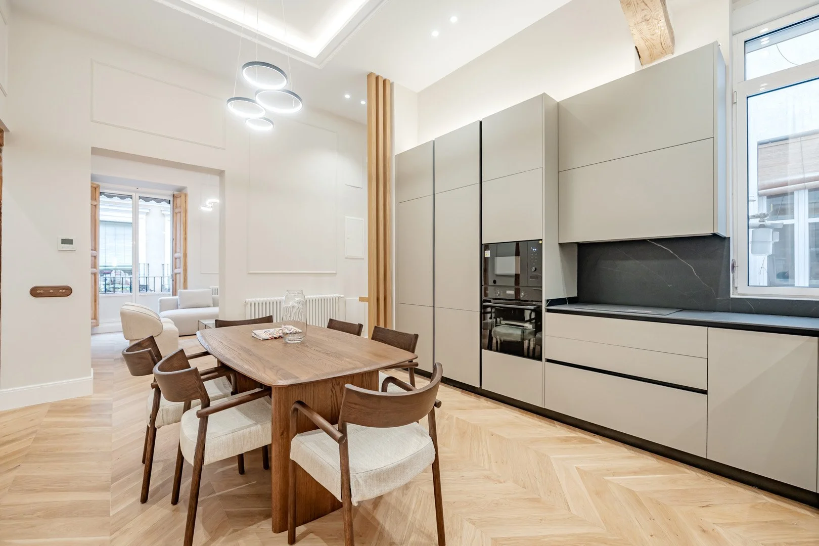 Modern kitchen and dining area with wooden table, six chairs, and sleek gray cabinetry, with a neighboring sunlit living space.
