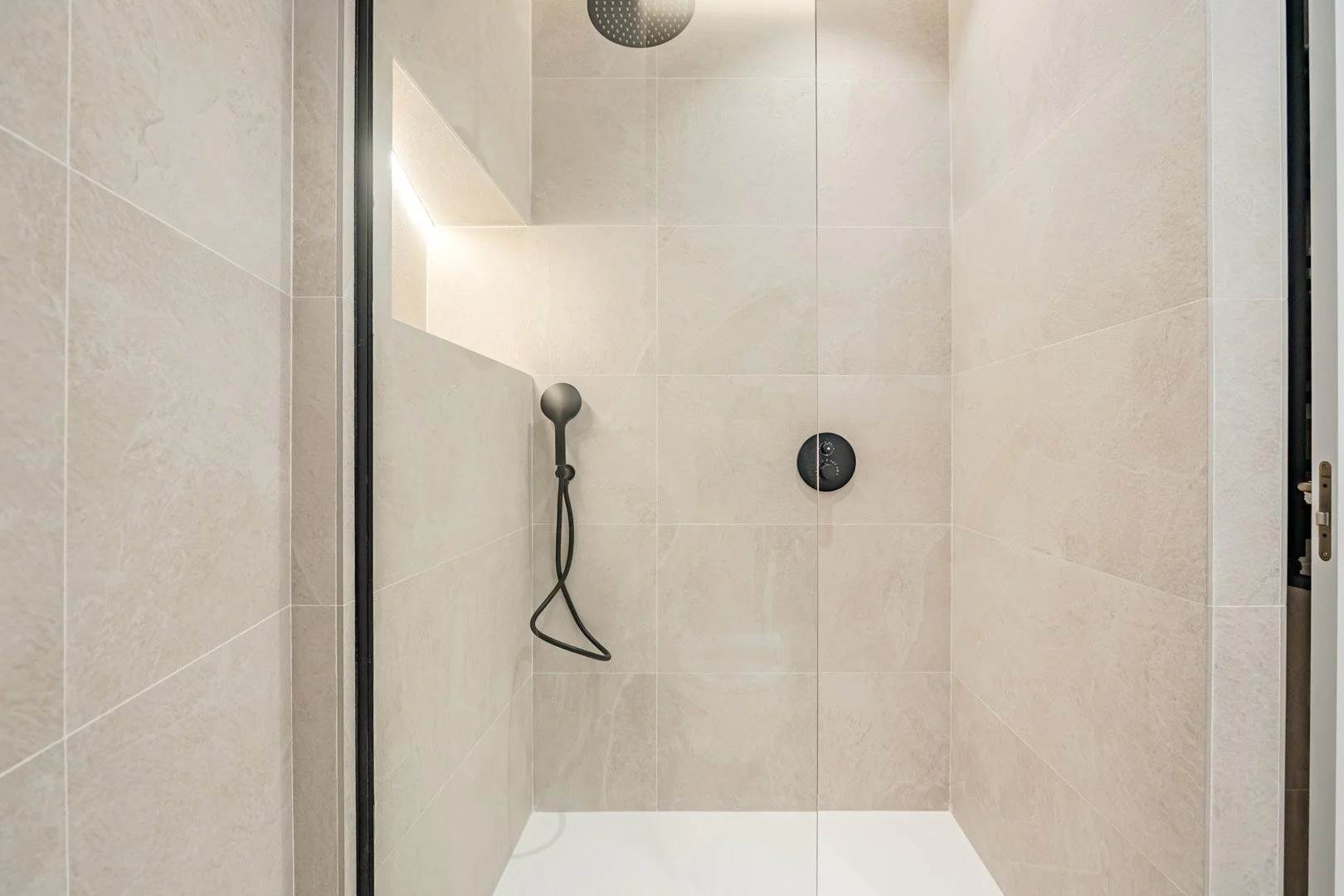 Empty shower stall with beige tiled walls, a black handheld showerhead, and a black control knob.