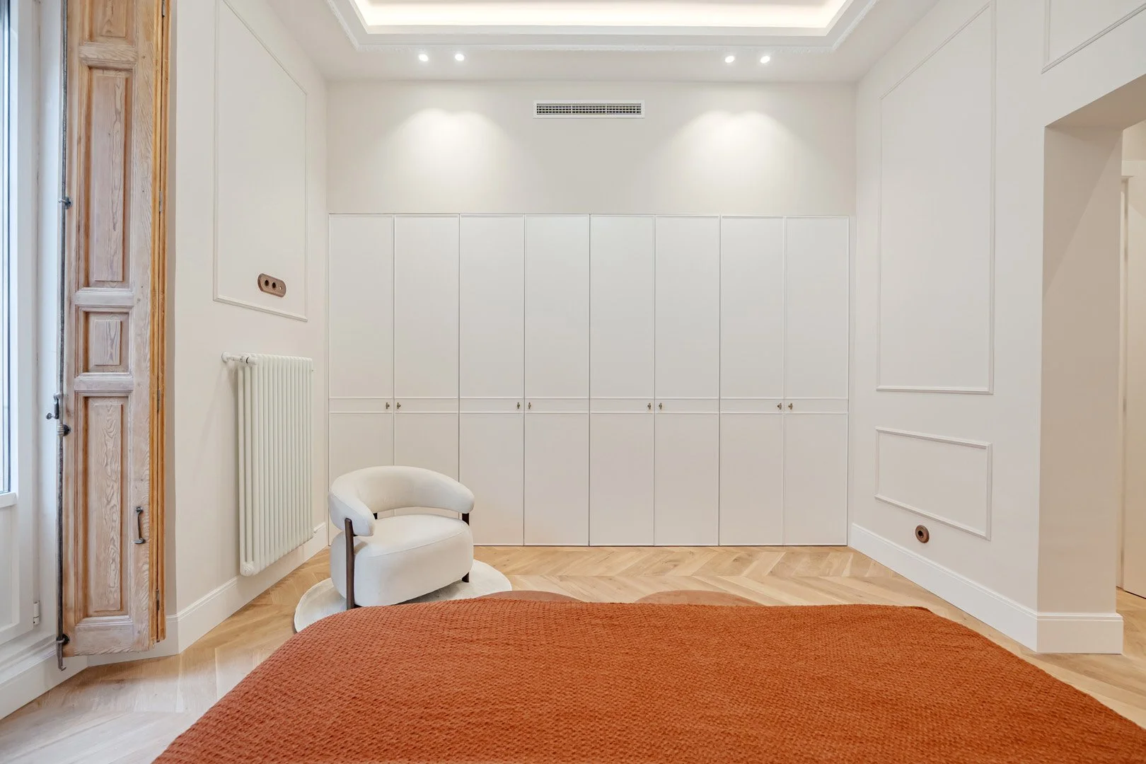 Side of a bedroom showing a large white closet, a round cream-colored chair on a small white rug, a wooden window with shutter, radiator, and part of a bed with an orange blanket.