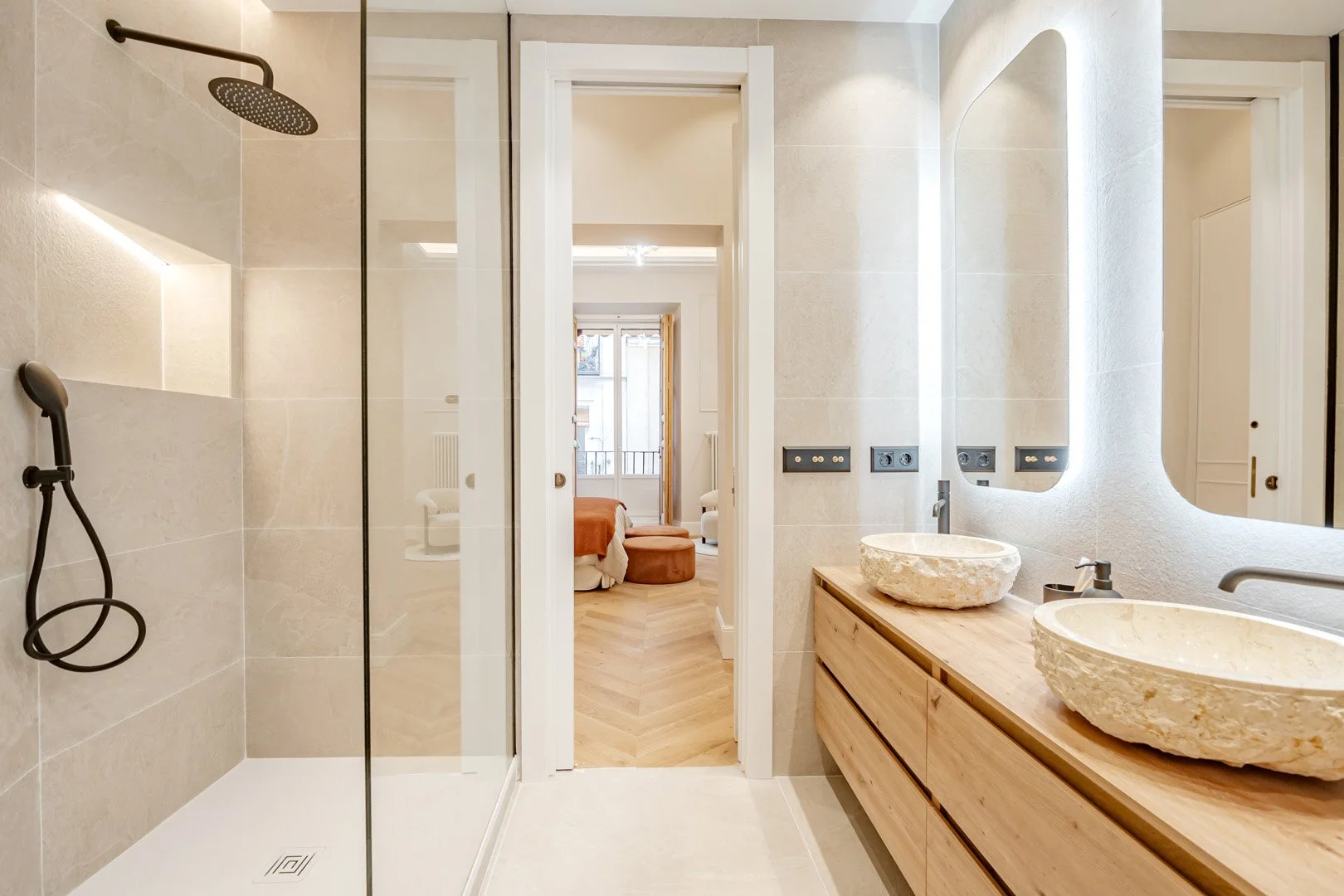 Modern bathroom with a walk-in shower, two stone sinks on a wooden vanity, large mirror with backlighting, and a view into a bedroom with a bed, a window, and a sitting area.