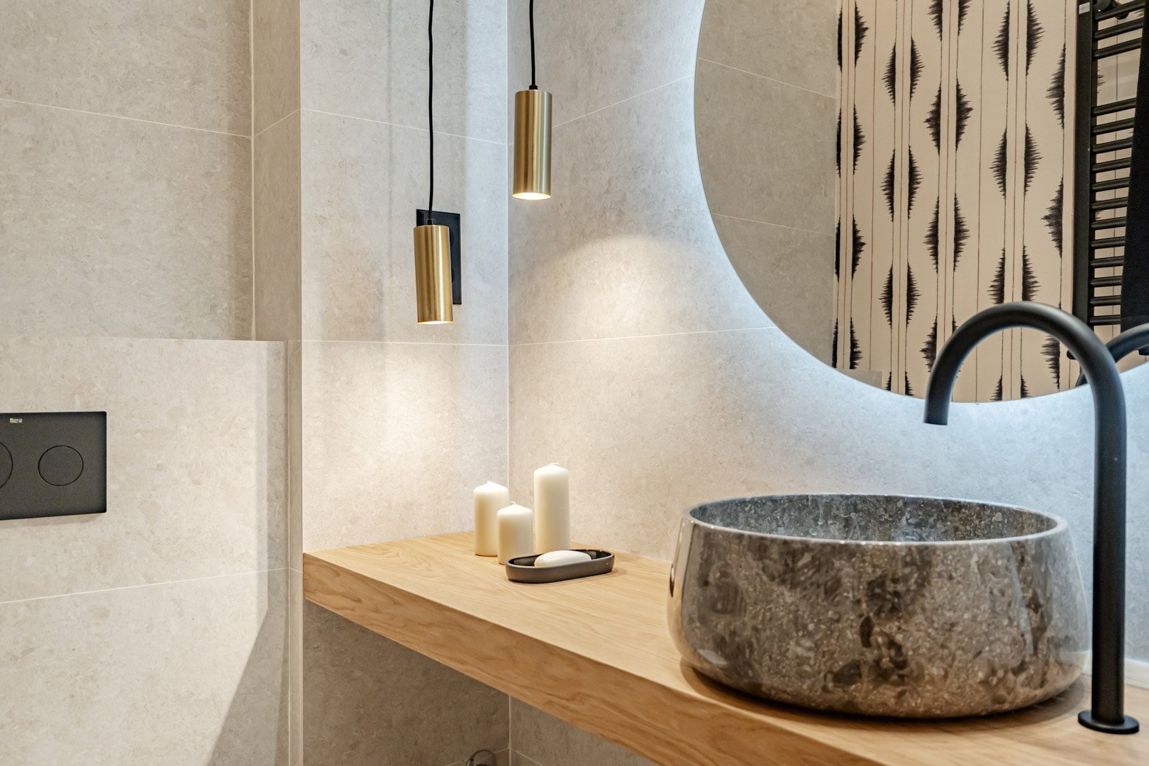 Modern bathroom with a round stone sink, black faucet, and wooden countertop, decorated with white candles, gold pendant lights, a large mirror, and patterned wallpaper.