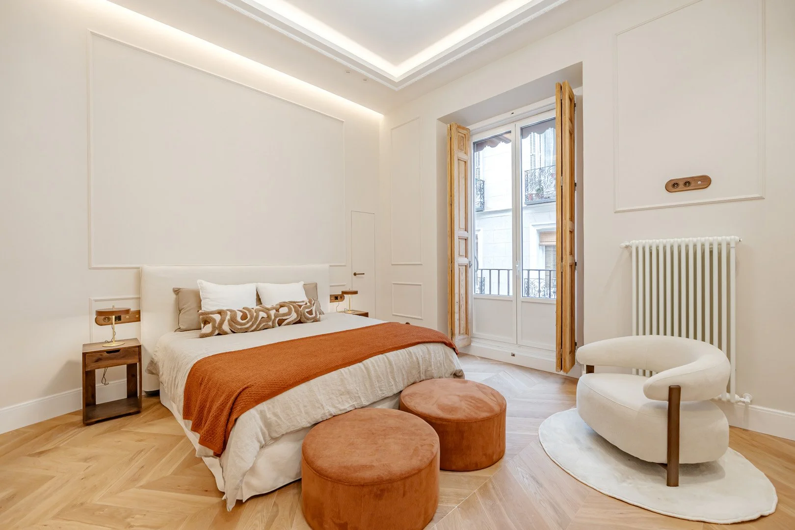 A minimalist bedroom with a white bed, brown and beige pillows, an orange blanket, two round orange ottomans, a unique white chair with wooden accents, a white rug, and a large window with wooden shutters.