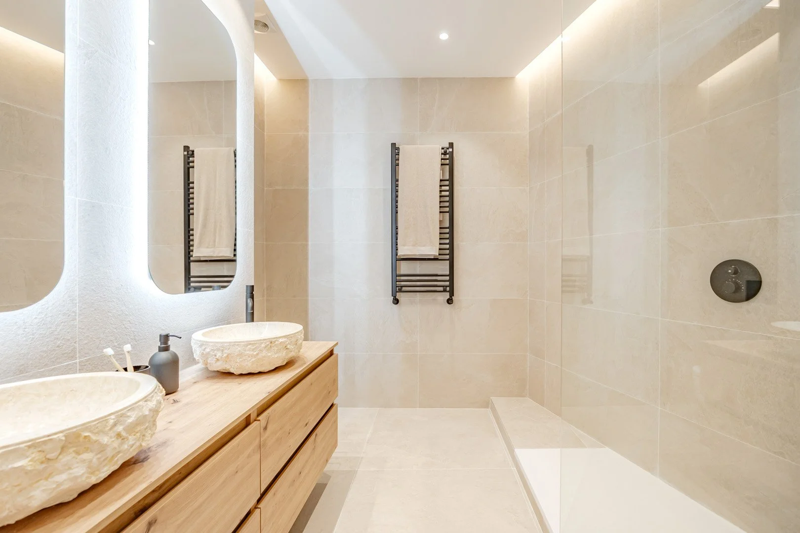 Modern bathroom with double stone vessel sinks on a wooden vanity, beige tiled walls and floor, two mirrors, towel rail with towels, and a walk-in shower area with a drain.