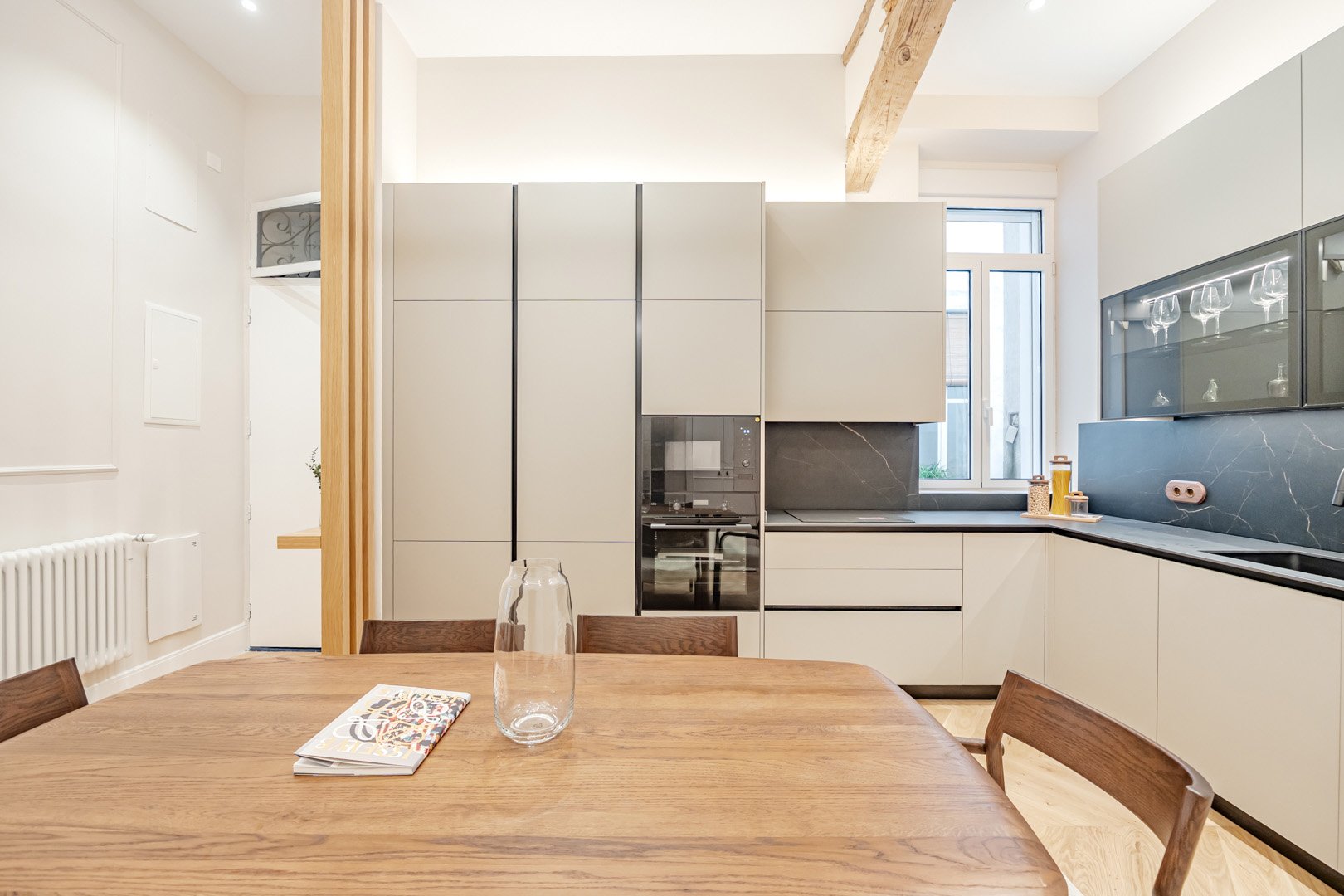 Modern kitchen with white cabinets, black countertops, a wooden dining table, and four chairs. There is a large window with natural light, a built-in oven, and a glass-front cabinet with glassware.