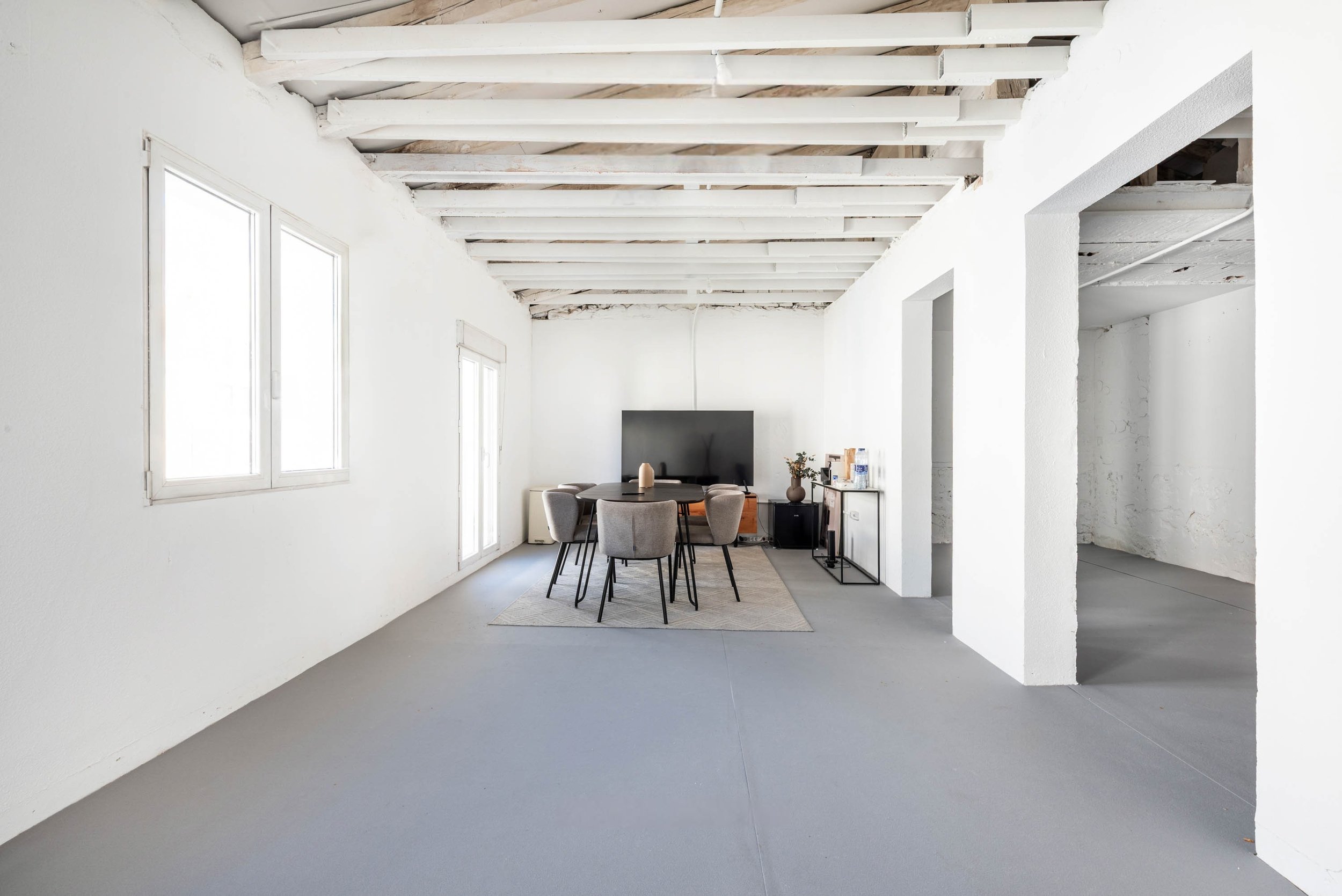 A minimalist living room with white walls, exposed wooden beams on the ceiling, two windows, a flat-screen TV mounted on the wall, a round dining table with six upholstered chairs, a light-colored rug, and a small black side table with decorative items and bottles.
