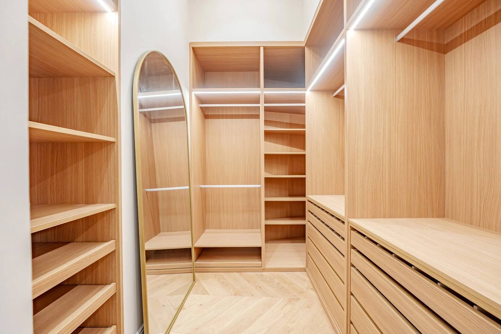 Empty walk-in closet with wooden shelves and drawers, a full-length mirror, and bright lighting.
