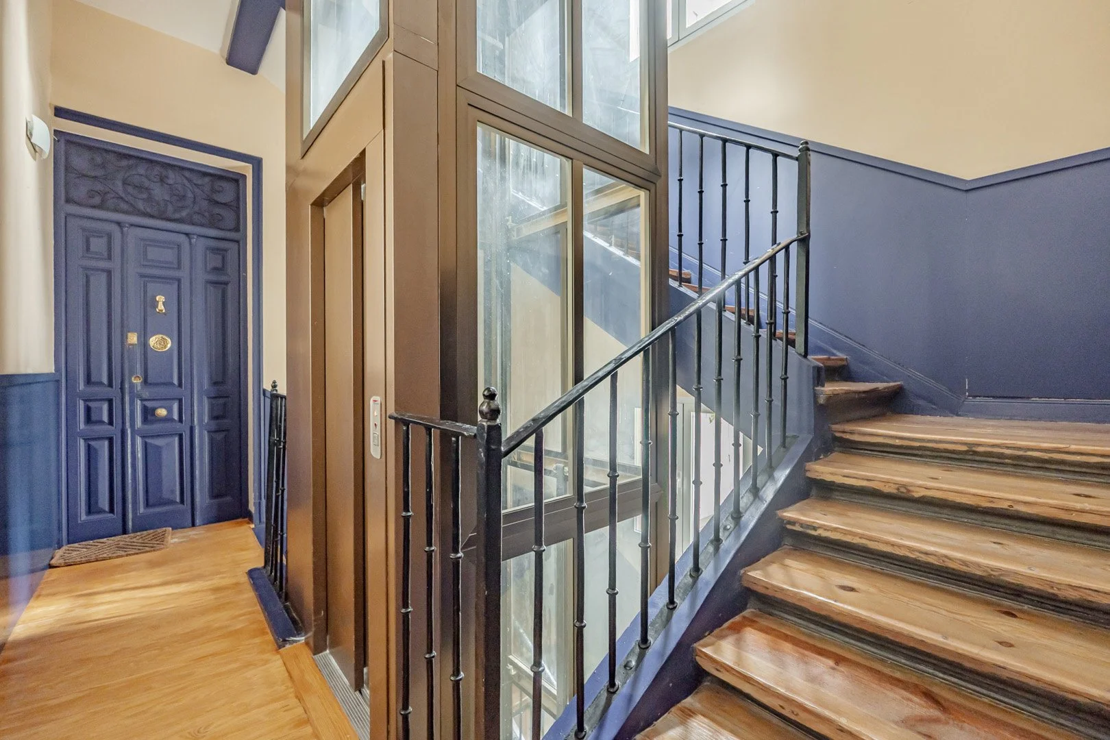 Interior staircase with wooden steps, black metal railing, blue walls, and a glass elevator shaft adjacent to the staircase.
