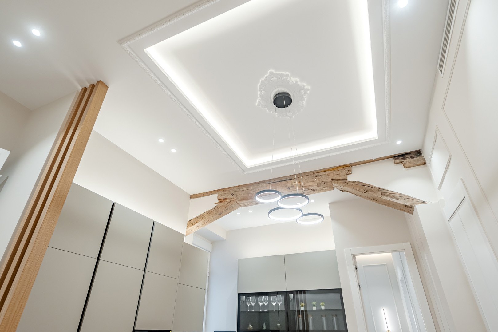 Modern interior ceiling with recessed lighting, decorative molding, exposed wooden beams, and a contemporary chandelier in a bright room.