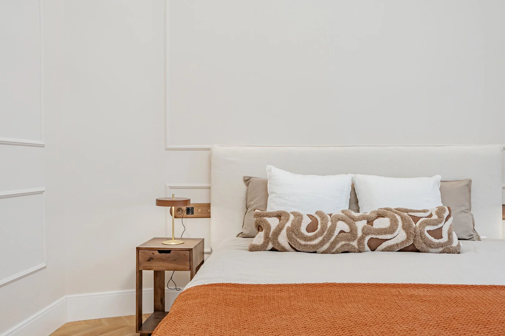A bedroom with a white bed, beige and white pillows, a brown and tan decorative pillow, a wooden nightstand with a small lamp, and a white wall with simple molding.