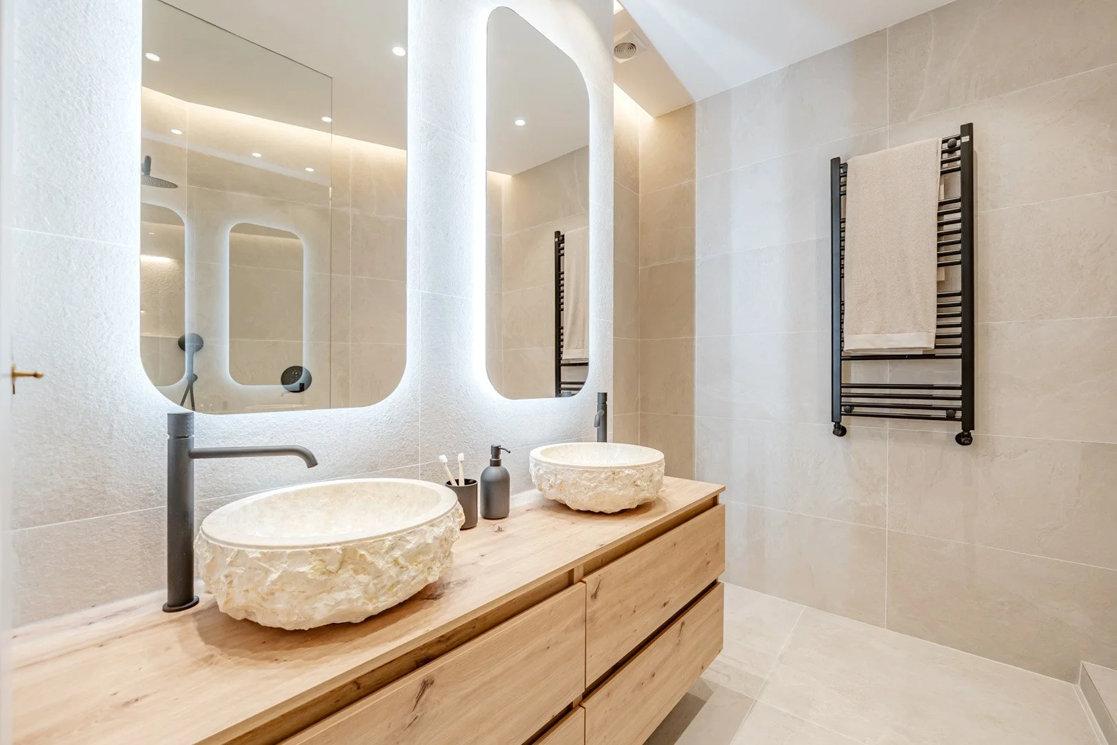 Modern bathroom with a floating wooden vanity, two stone vessel sinks, large wall mirror, black towel radiator, beige tiled walls and floor, black soap dispenser, and toothbrush holder.