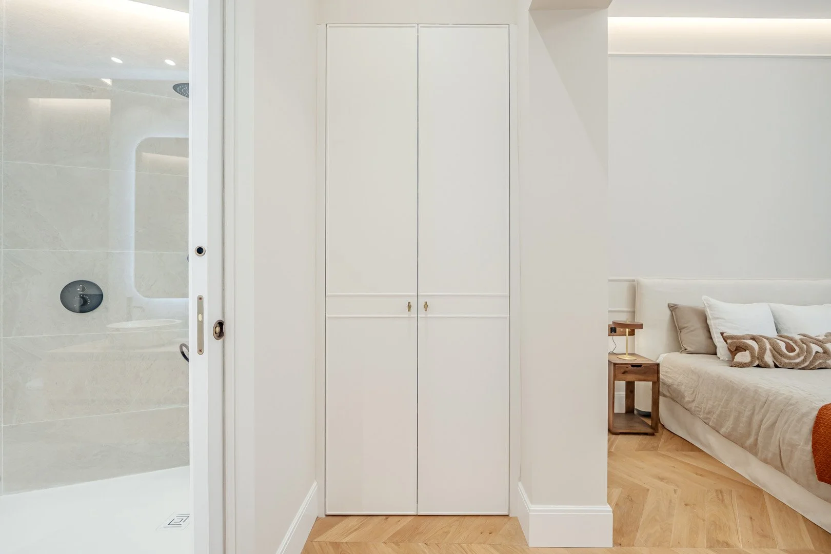 A modern bedroom with a white bed, beige and white pillows, a wooden nightstand with a small lamp, a wardrobe, and an en-suite bathroom with a walk-in shower seen through a sliding glass door.
