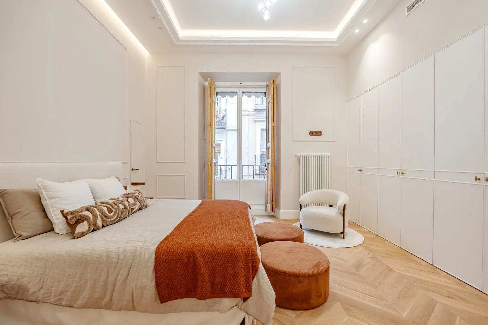 A bedroom with a large bed, a window with wooden shutters, a small white chair, two round orange ottomans, a white oval rug, and built-in white cabinets.