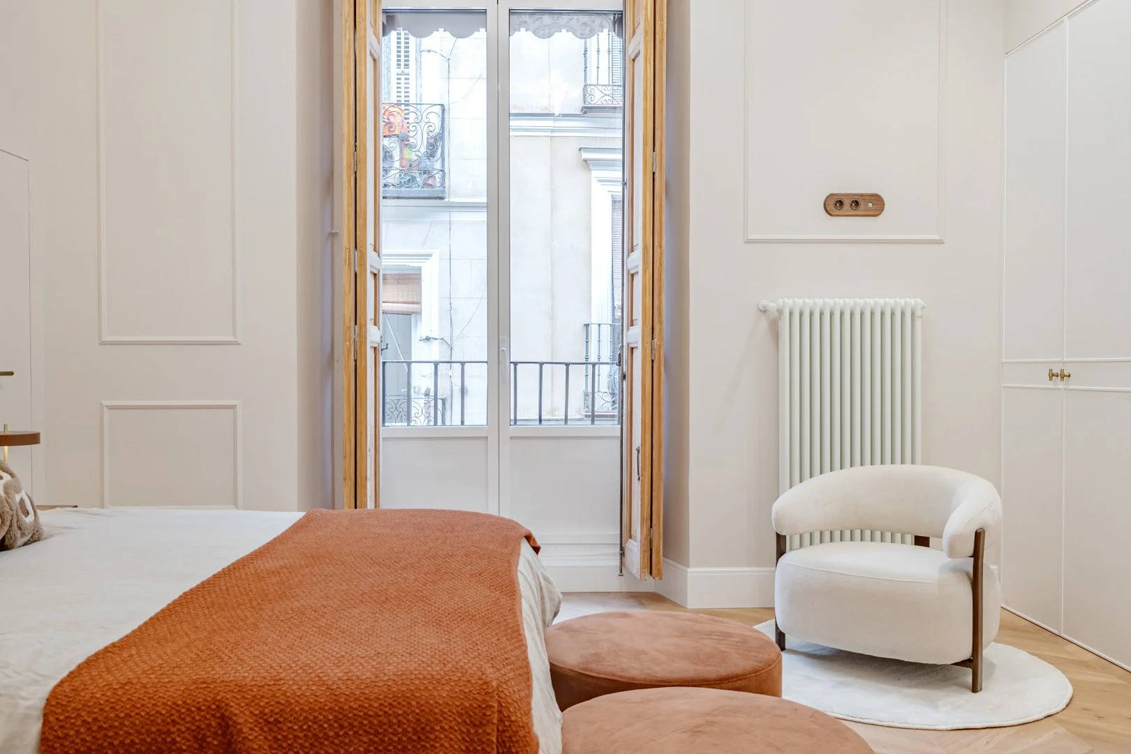 A bedroom with a white bed covered with an orange blanket, a pair of round ottomans, and a white armchair in front of a window with French doors leading to a balcony.