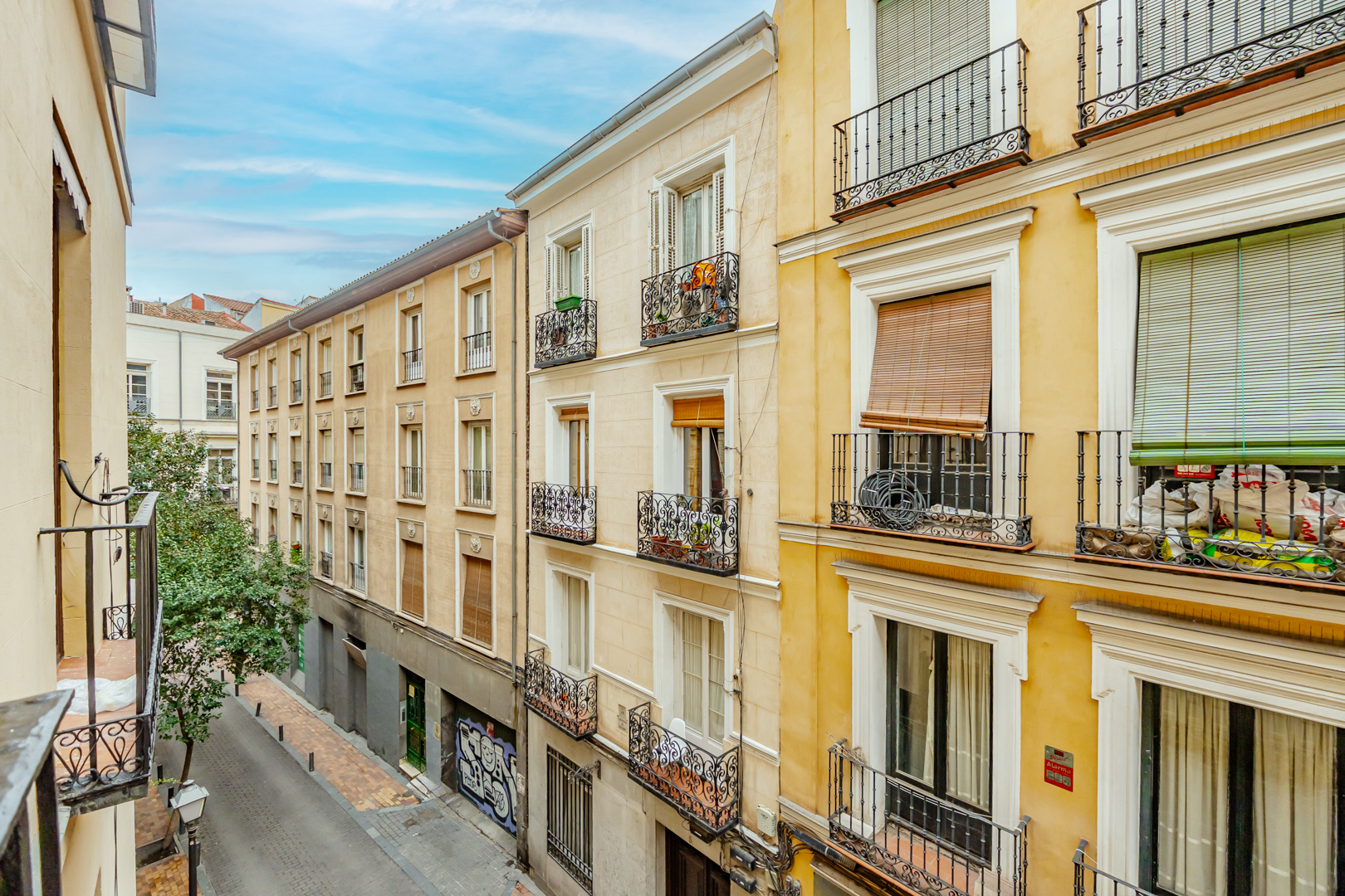 View of a street with yellow and beige apartment buildings, some with balconies and window shutters, and a tree on the sidewalk, under a partly cloudy sky.