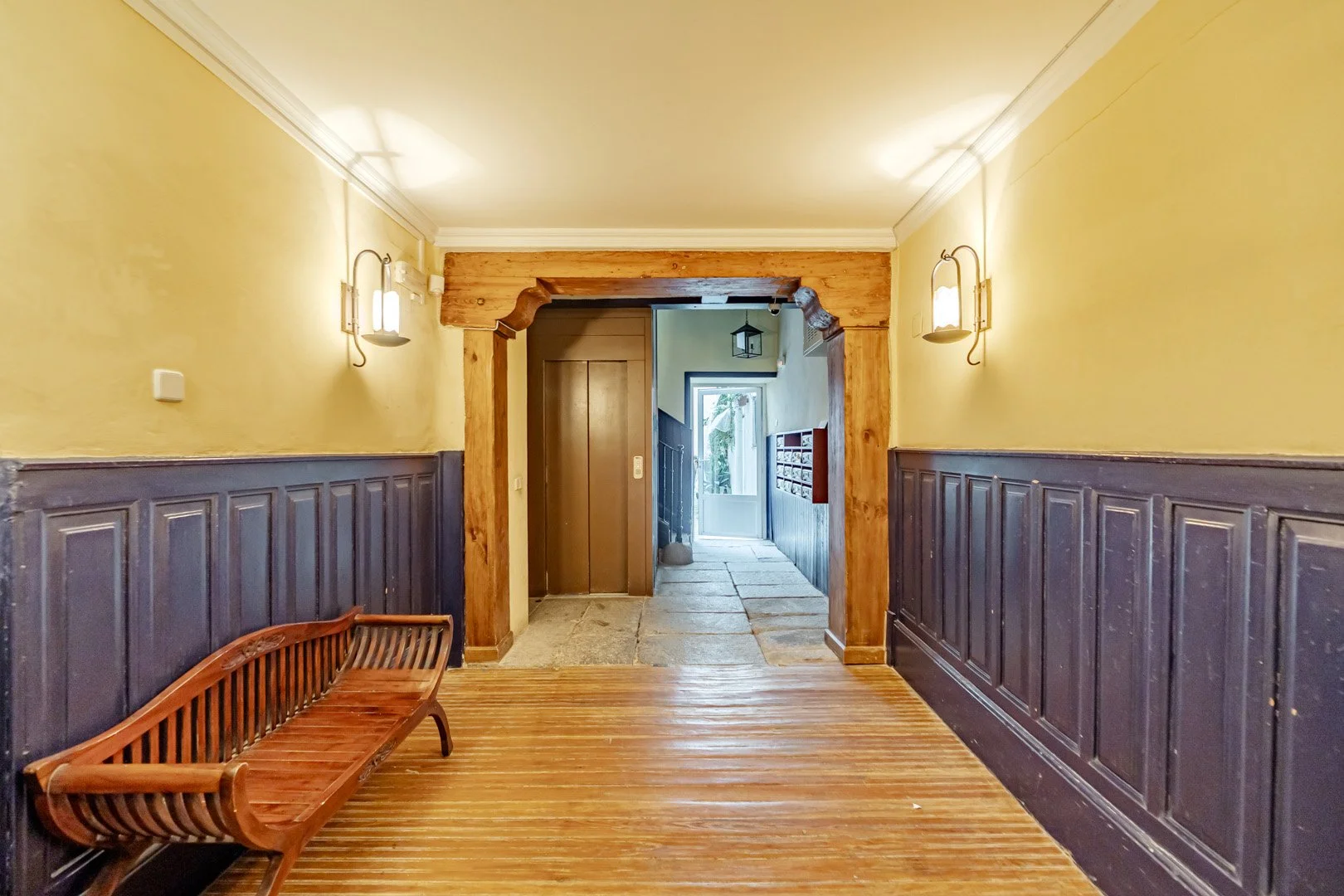 Hallway with yellow walls, dark wood wainscoting, wooden bench on the left, and wall-mounted lights. At the end, a doorway, window, and staircase are visible.