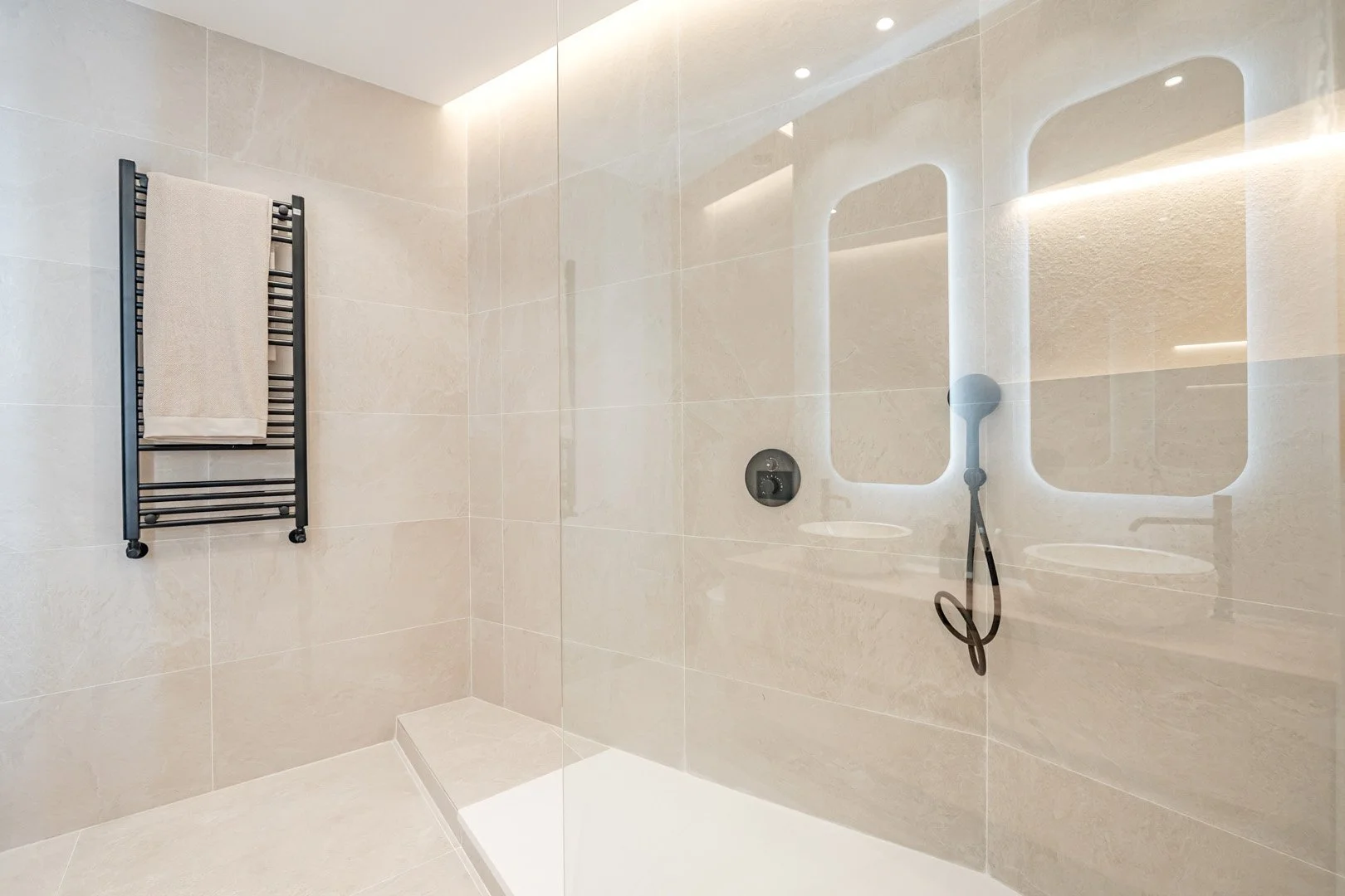 Modern, minimalist shower with beige tiled walls and floor, black towel radiator with a towel, and a round showerhead with a black hose and control panel.
