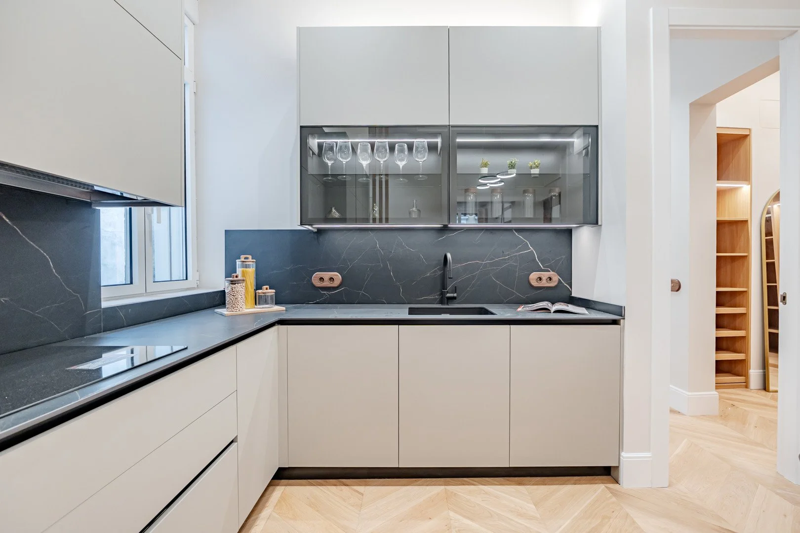 Modern kitchen with white cabinets, a black marble countertop and backsplash, black faucet, and glass-front upper cabinets containing glasses and small plants. There are three electrical outlets and a window allowing natural light.