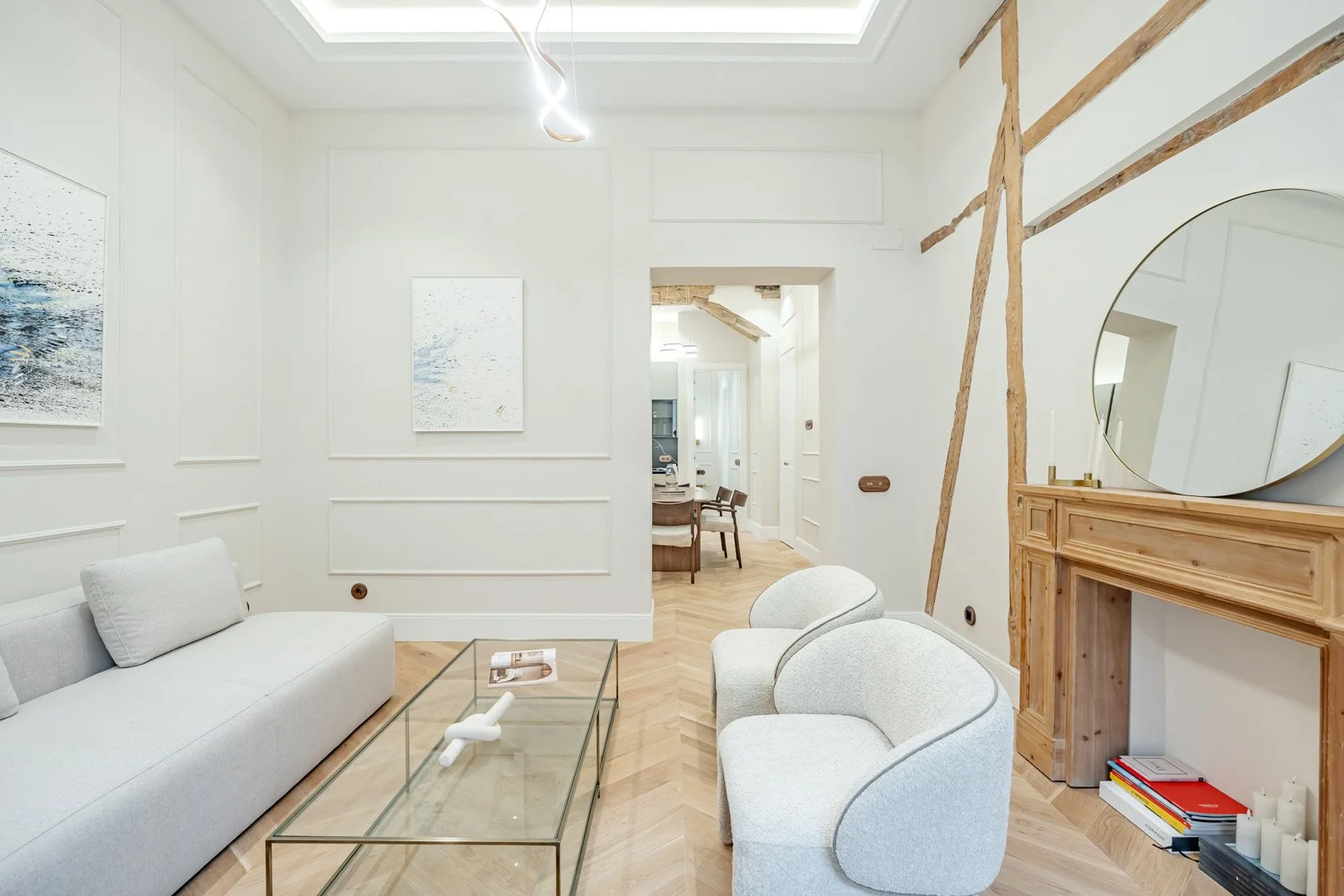 A modern, minimalist living room with white walls, light wood flooring, and simple furniture, including a white sofa, two white armchairs, a glass coffee table, and a wooden fireplace mantel with a round mirror above it. There are artworks on the wal