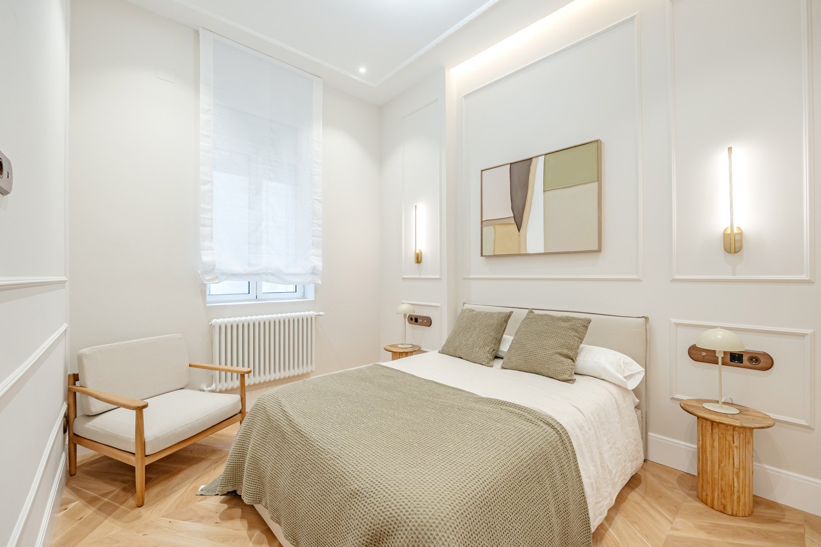 A minimalist bedroom with a white bed, beige pillows and blanket, wooden side tables with lamps, a white armchair with wooden arms, a window with sheer white curtain, and framed abstract art on the wall.