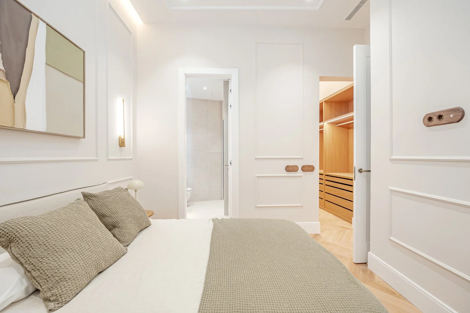 A bedroom with a bed, beige pillows, white bedding, wall art, a wall-mounted light, a wooden nightstand, and a doorway leading to a bathroom and a closet with wooden shelving.