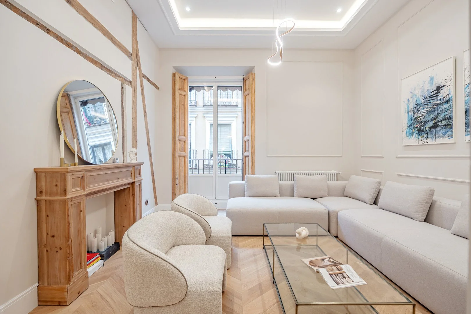 A modern living room with a white sectional sofa, a glass coffee table, and two curved beige chairs. There is a wooden fireplace mantel with candles and books, and abstract artwork on the wall. A large window with wooden shutters opens to a balcony w