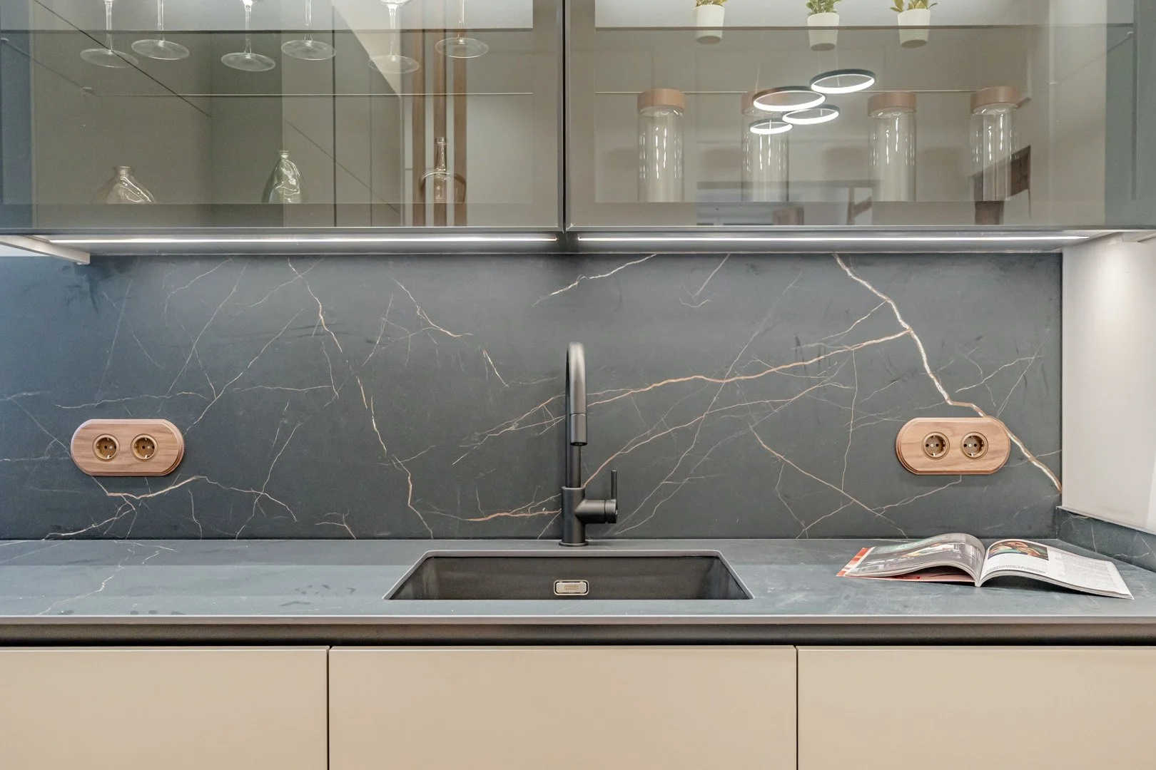 Modern kitchen sink area with black marble backsplash, a faucet, and two electrical outlets with wooden covers. A magazine is open on the right side of the counter above white cabinets. Glass cabinets with various decorative items are above.