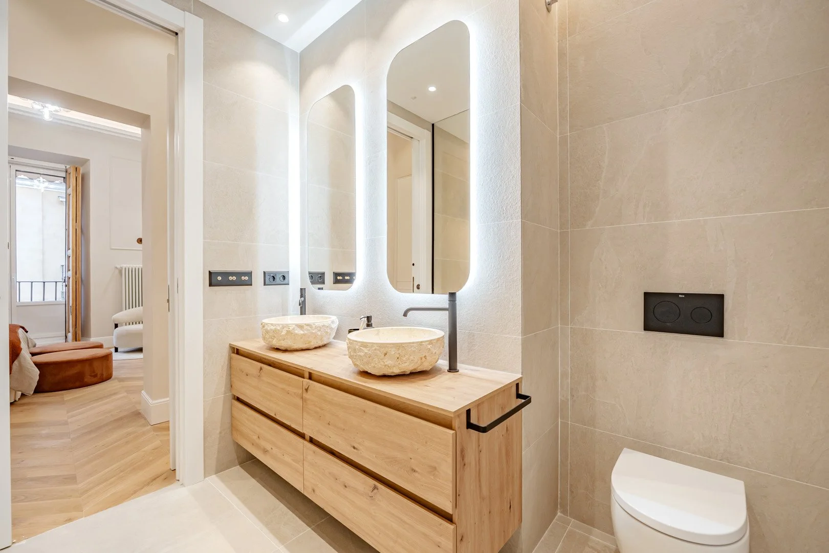 Modern bathroom with dual stone vessel sinks, large mirrors with built-in lighting, wooden vanity, beige tile walls, and a wall-mounted toilet.