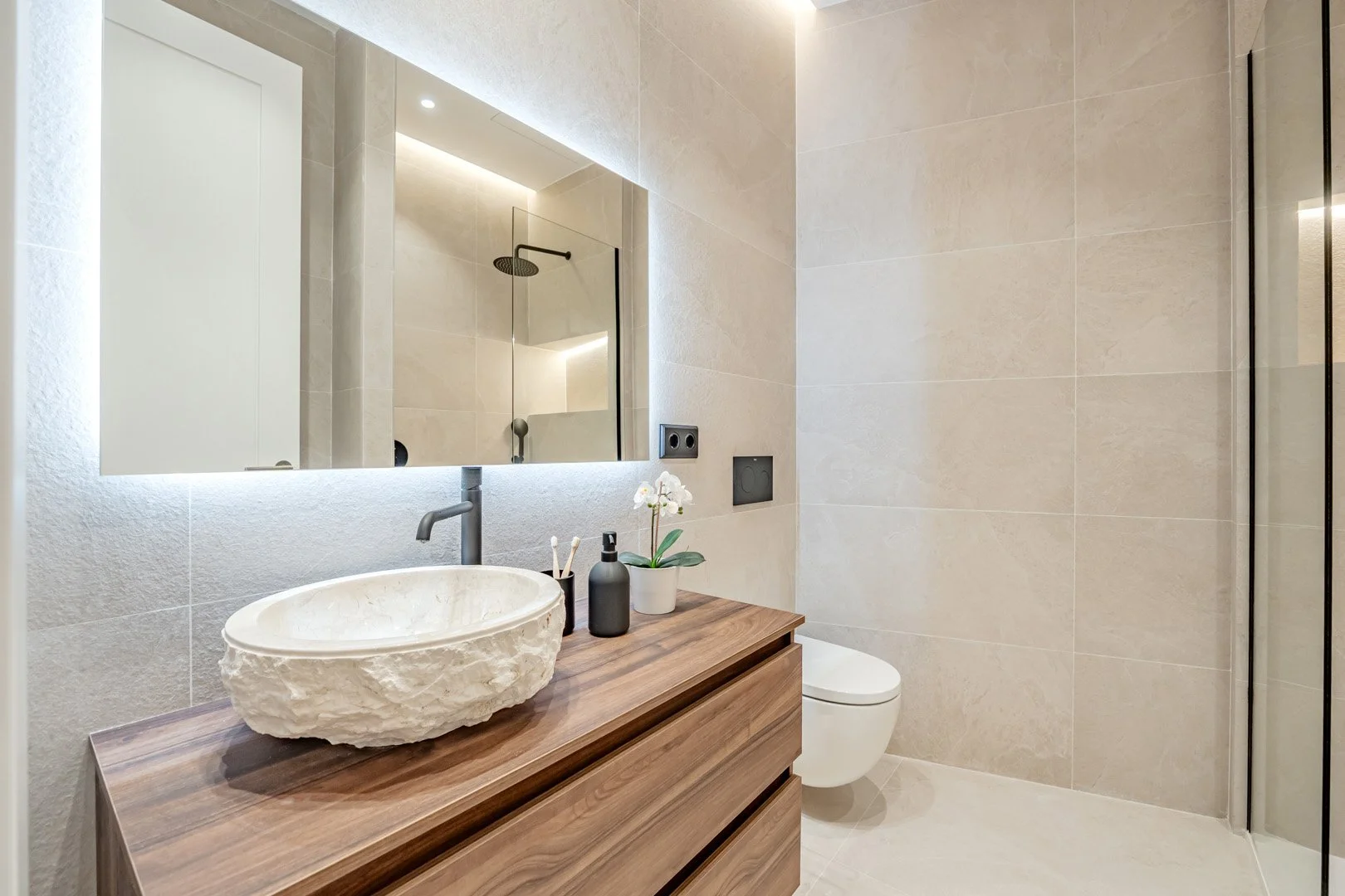 Modern bathroom with beige tiled walls, a wooden vanity with a textured stone sink, a large mirror with backlighting, a small potted orchid, soap dispenser, and toothbrush holder, a wall-mounted toilet, and a walk-in shower with a glass door and rain