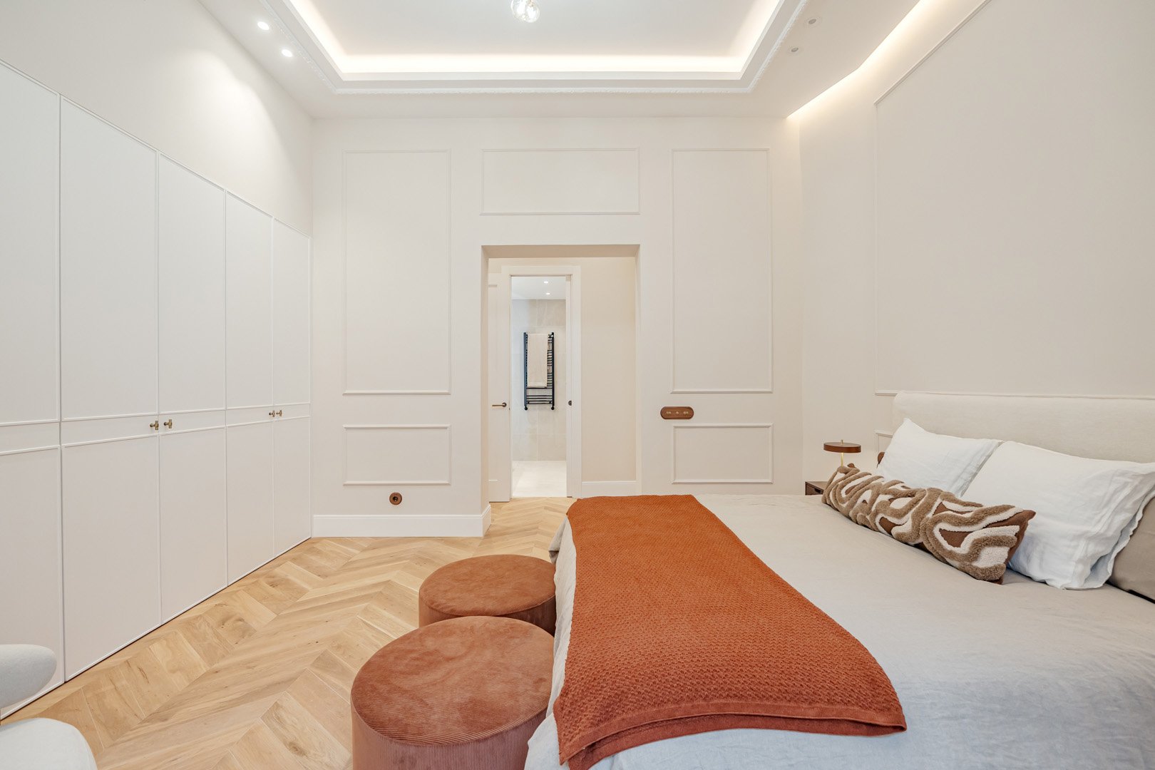 A minimalist bedroom with white walls, a white bed with pillows, a rust-colored blanket, and two round rust-colored stools. There is a wooden side table, a large white wardrobe, and a doorway leading to a bathroom with towels on a rack.