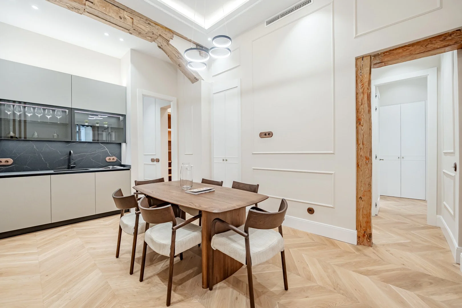 Modern dining room with a wooden table surrounded by six chairs, a built-in kitchen with black countertops and gray cabinets, and wooden accents on the ceiling beam and doorway frame.