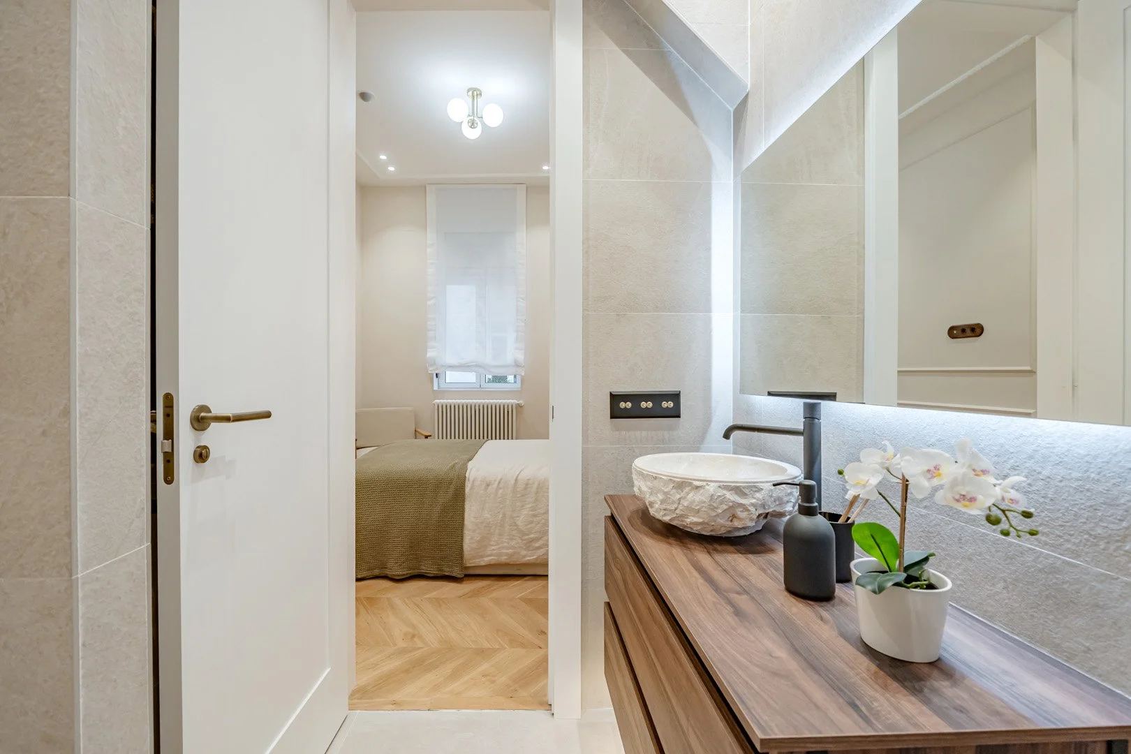Modern bedroom visible from bathroom area with beige bed, window with sheer white curtains, and radiator. Bathroom vanity with stone sink, wooden countertop, black faucet, black bottle, and orchid plant, mirror reflecting white wall.