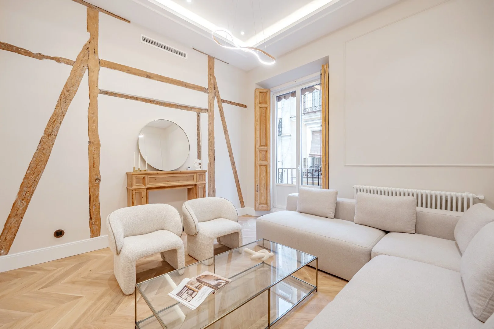 Modern living room with cream sofa, two cream armchairs, glass coffee table, decorative wood beams on the wall, round mirror above fireplace, large window with wooden shutters, and minimalist lighting fixture.